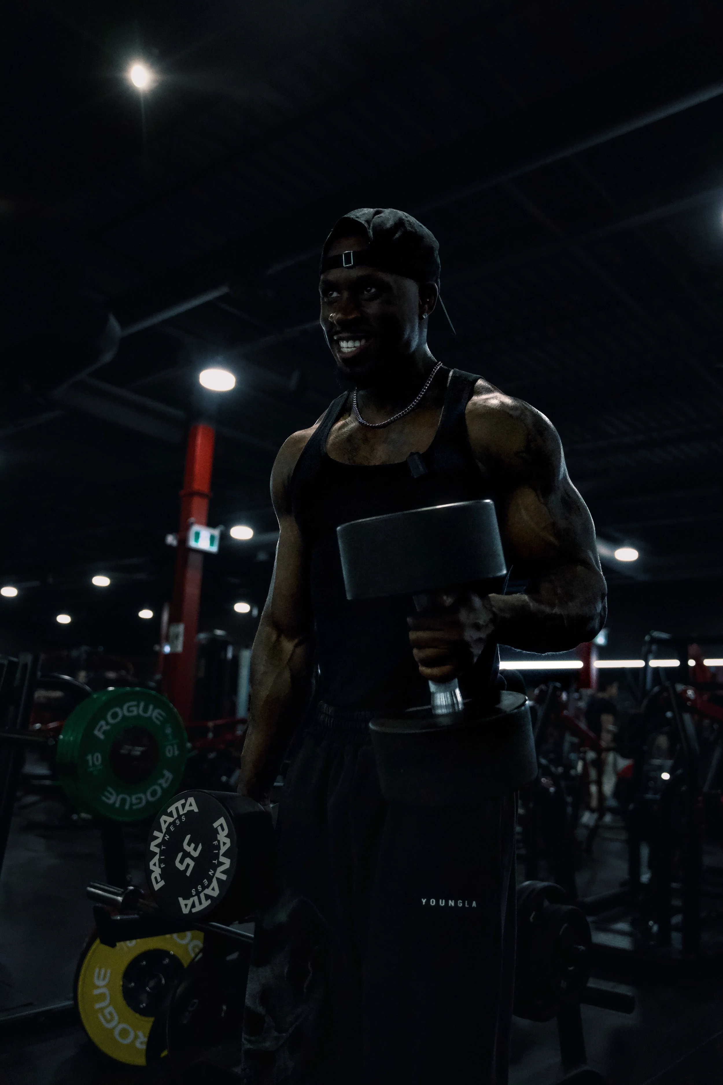A muscular man wearing a black cap, tank top, and black pants, smiling while holding a dumbbell in each hand in a dark gym setting with bright spotlights.