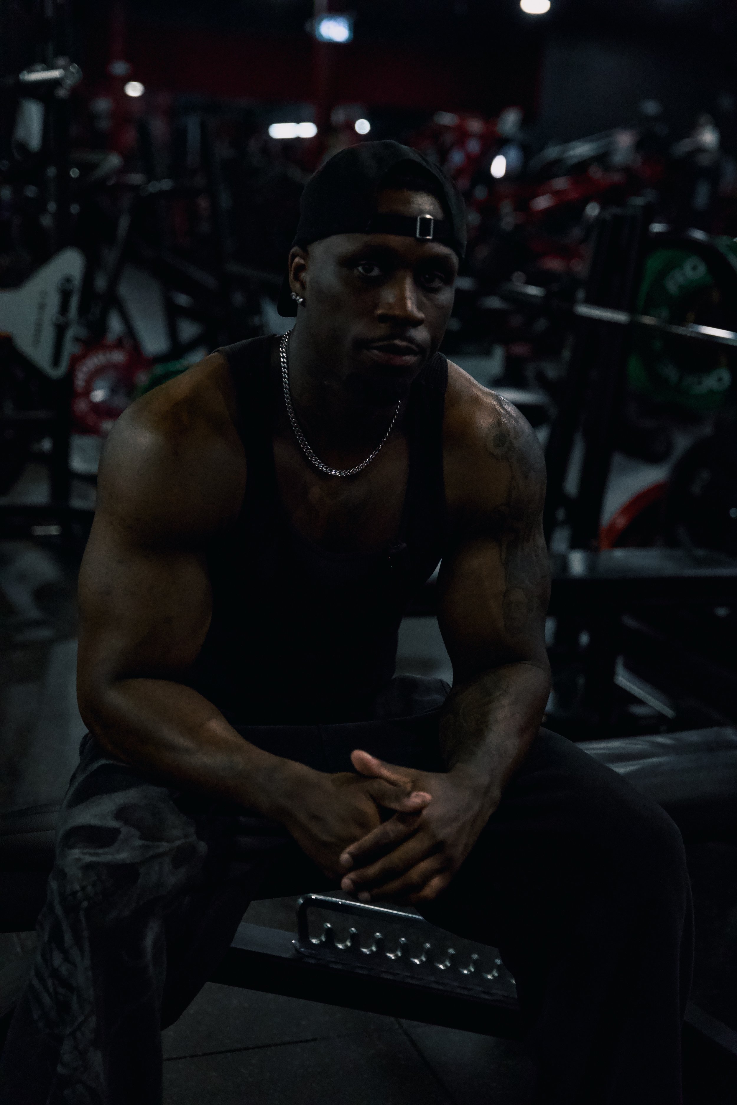 A muscular man wearing a black tank top, black cap backwards, and silver chain necklace, sitting in a gym with weightlifting equipment in the background.