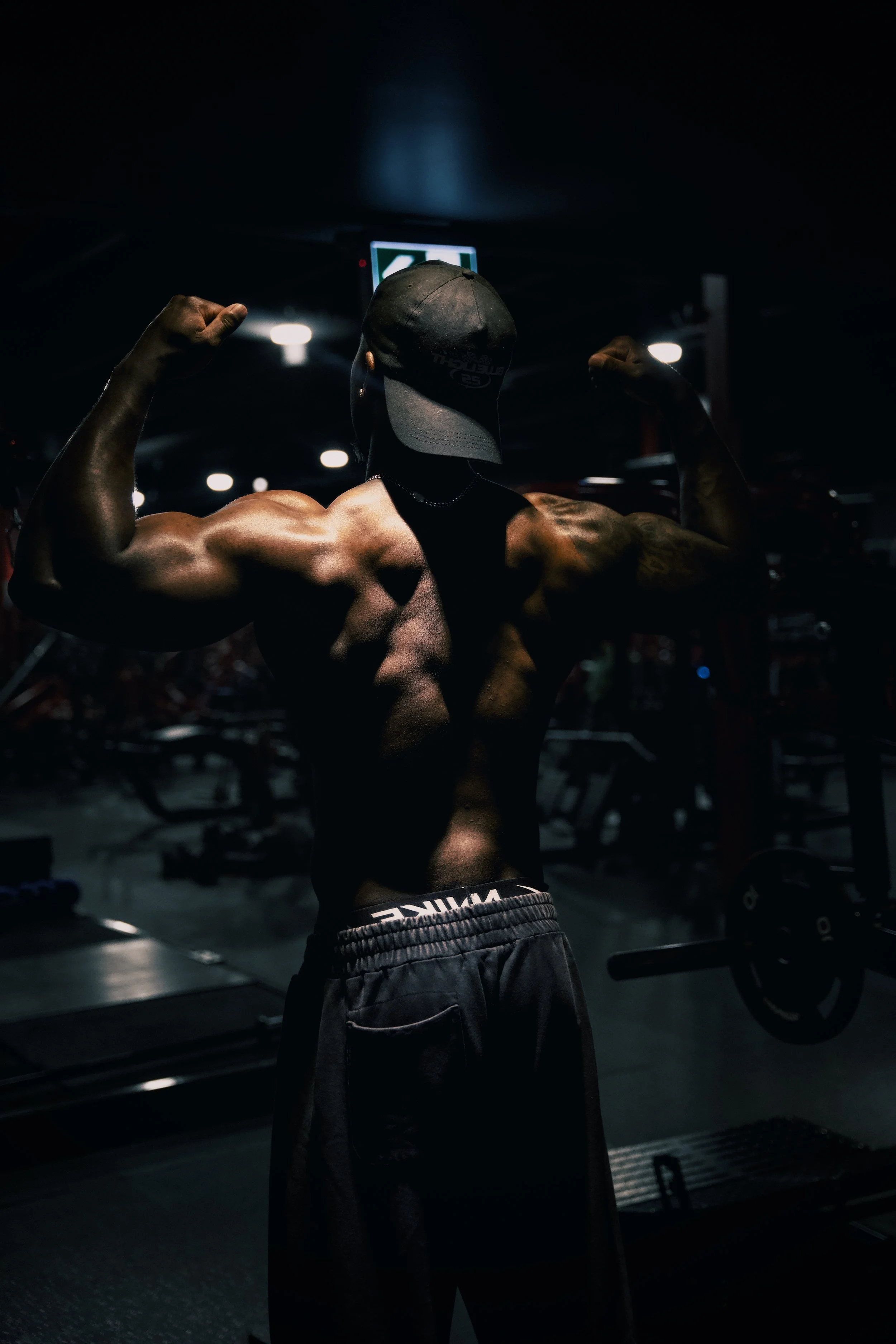 A muscular man in a dark gym, wearing a cap and dark shorts, flexing his arms with weightlifting equipment in the background.