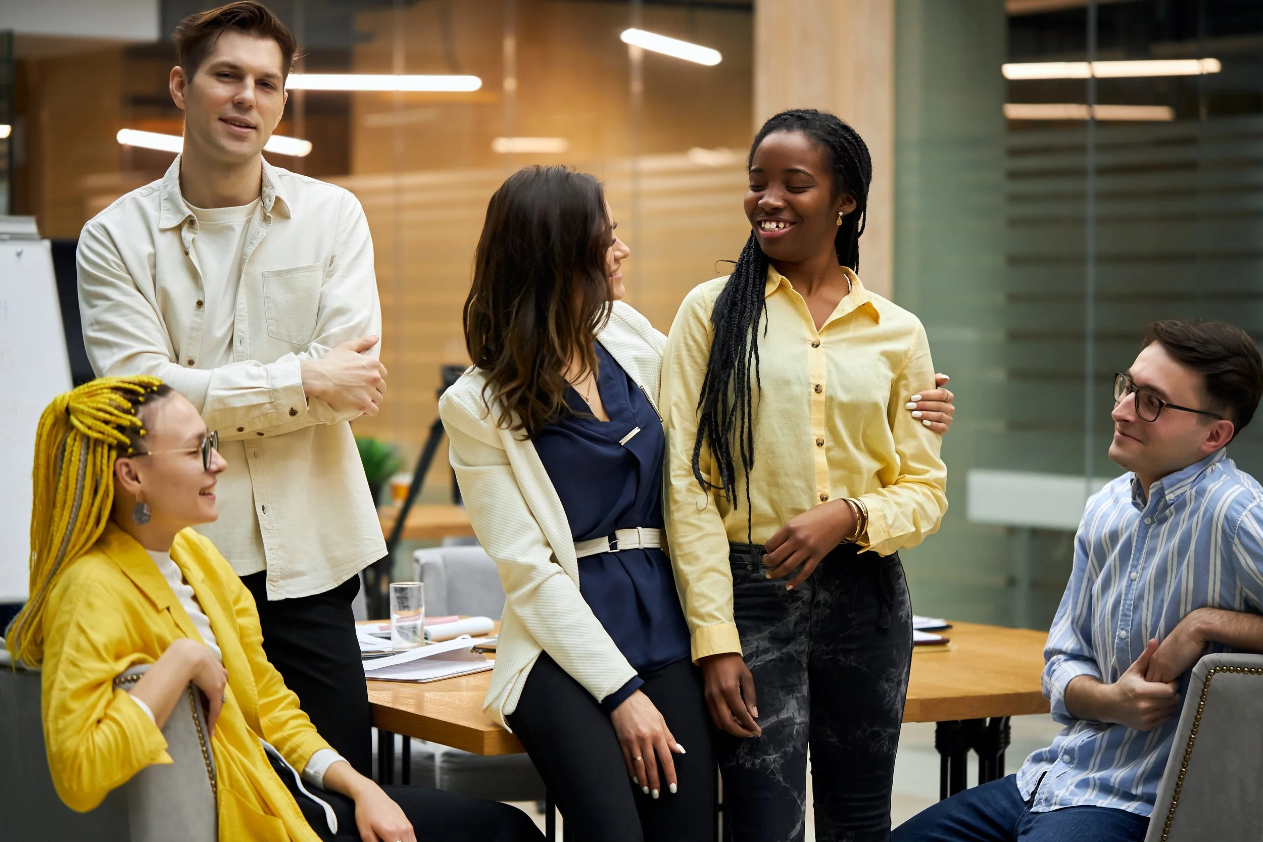 A diverse group of people in a modern office, with three women and two men, engaging in conversation and smiling.