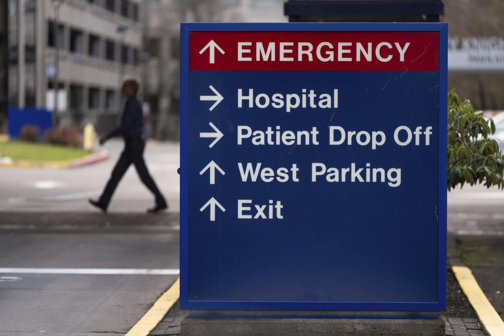 Hospital directional sign with white text on a blue background, indicating routes to emergency, hospital, patient drop off, west parking, and exit, with a pedestrian walking past in the background.