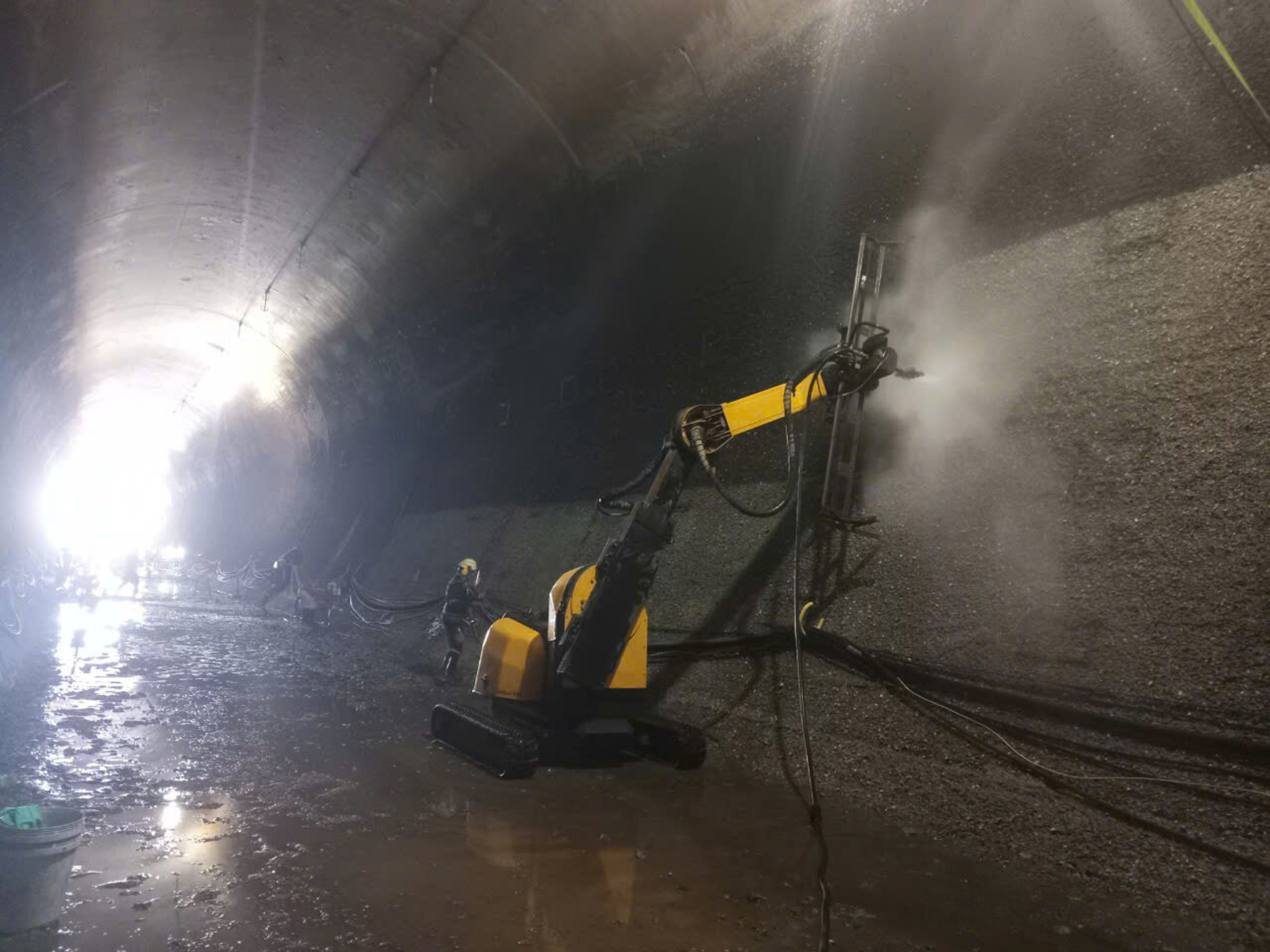Industrial tunnel construction or maintenance with a yellow robotic machine spraying or cleaning the tunnel wall while a worker observes nearby.