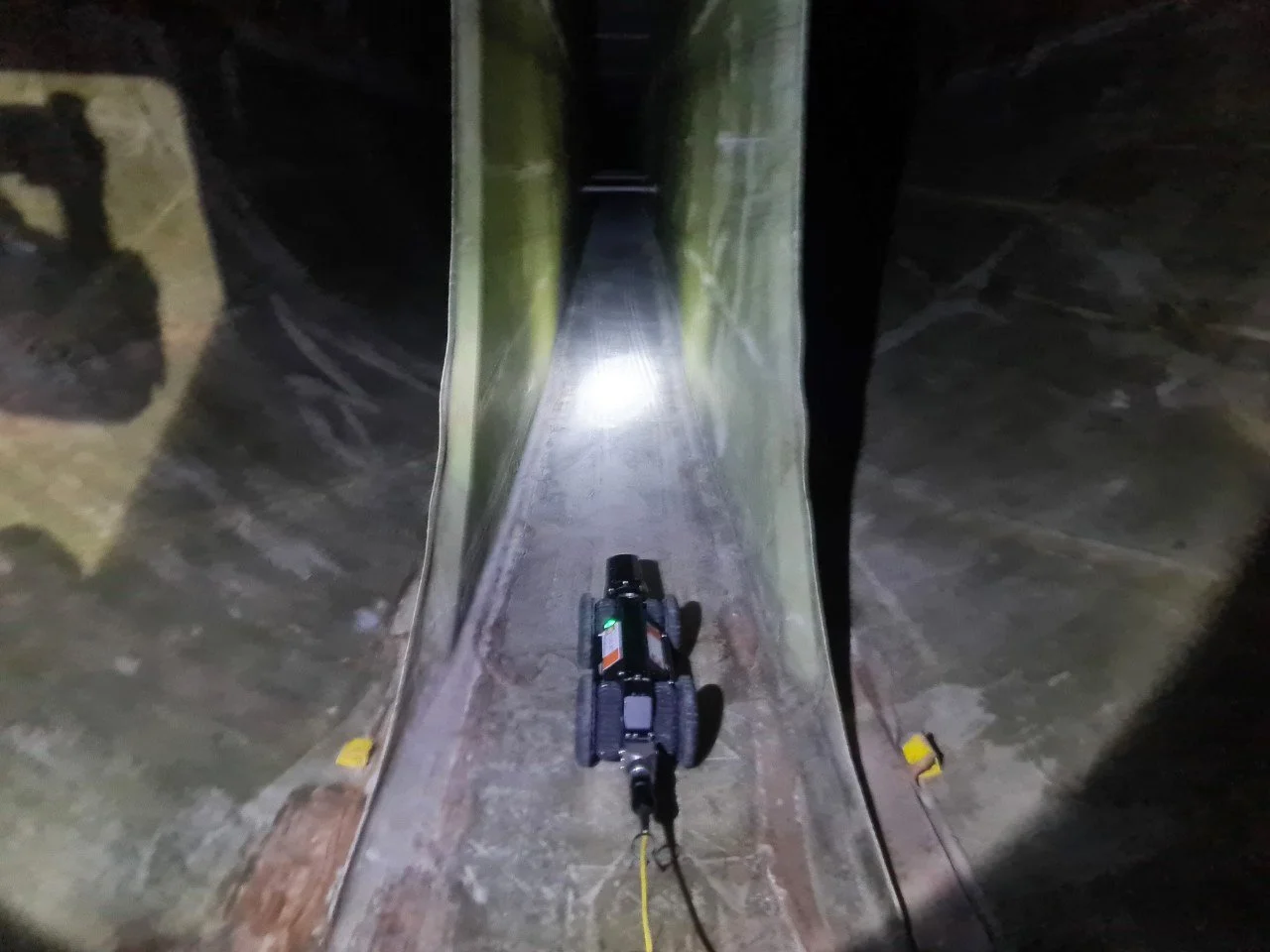 A mirror maze with walls made of large reflections of the surrounding area, and a flashlight illuminating the pathway. A small robot or device is on the ground in the center of the passage.