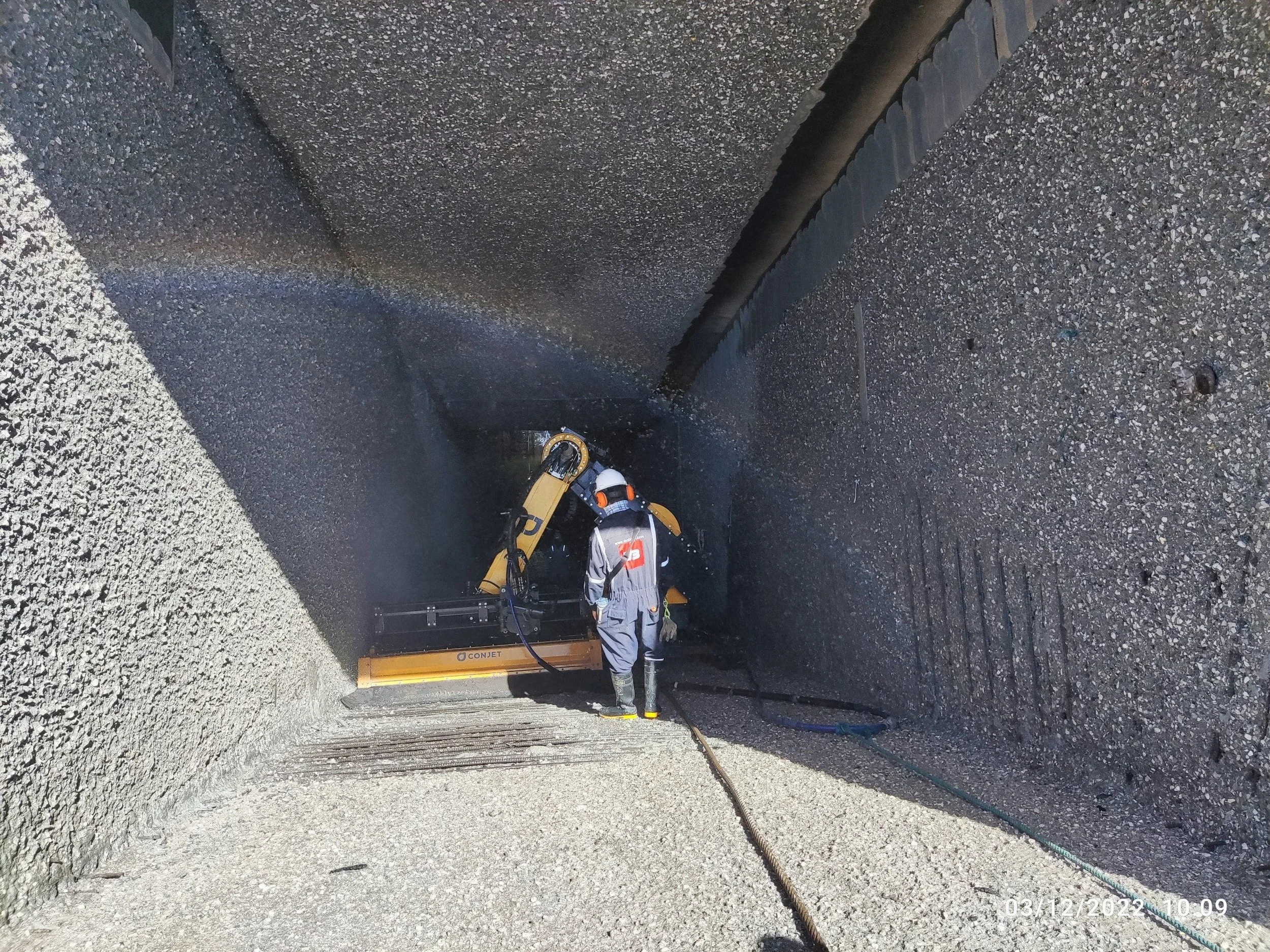A worker in protective gear operating a machine inside a large, deep, concrete-lined tunnel or trench.