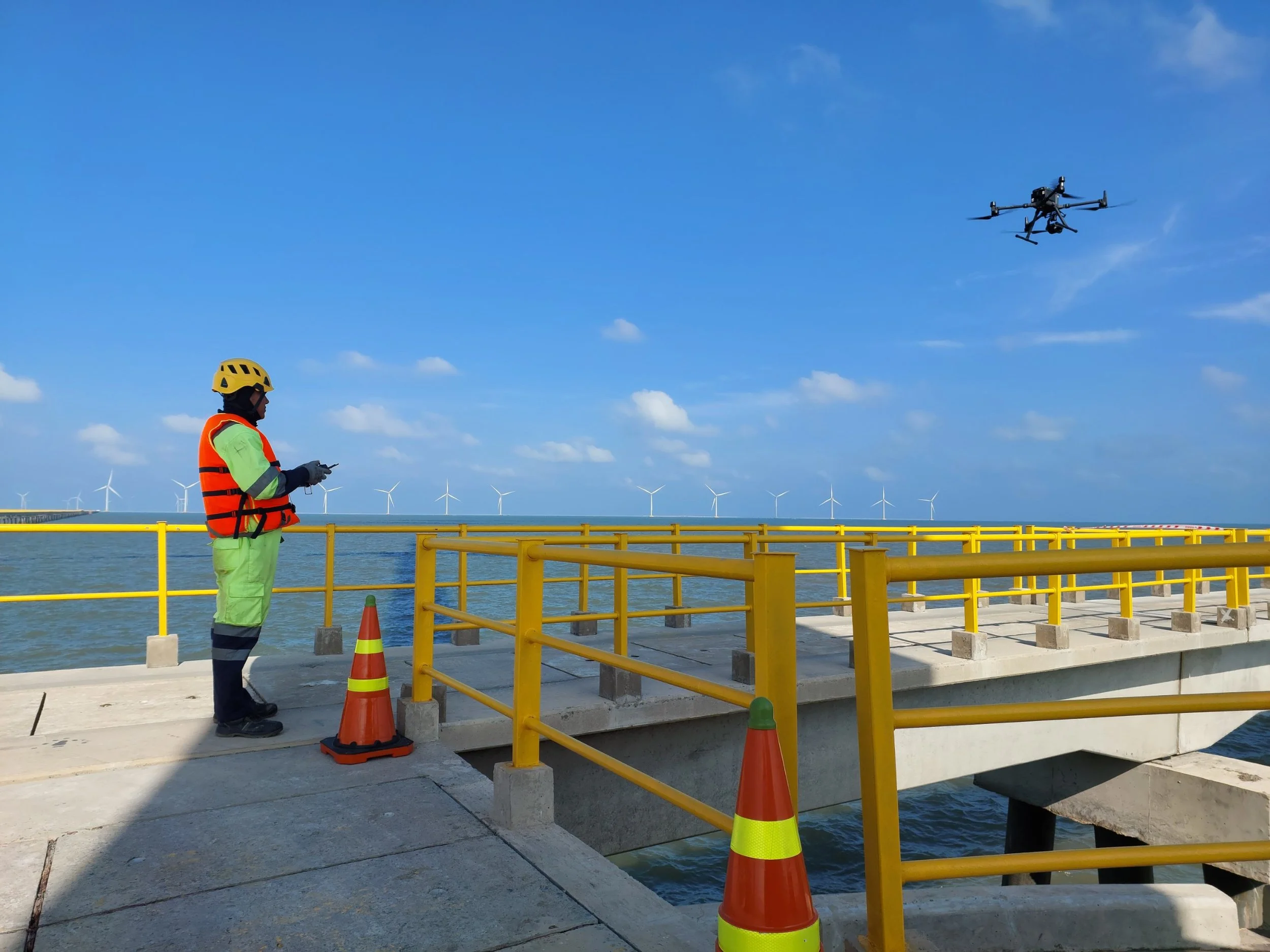 Person in safety gear flying a drone over an offshore platform with wind turbines in the background.