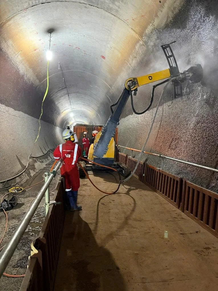 Workers in protective gear using specialized equipment to drill a large underground tunnel, with concrete walls and safety barriers visible.