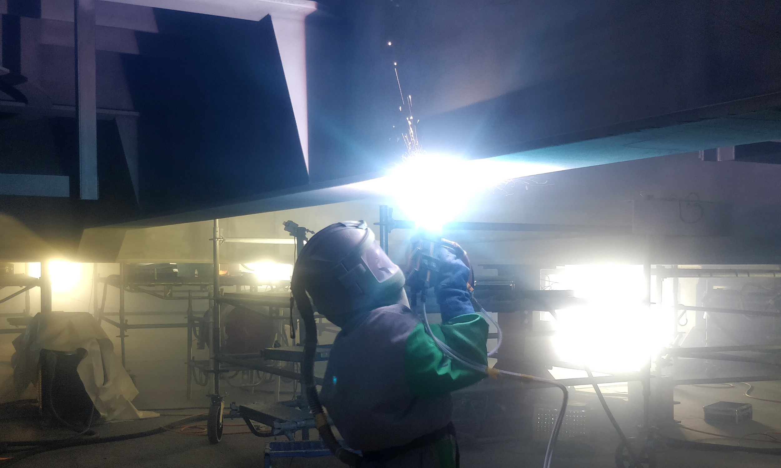 A worker in safety gear welding metal beams in an industrial setting, with bright sparks and shielded face.