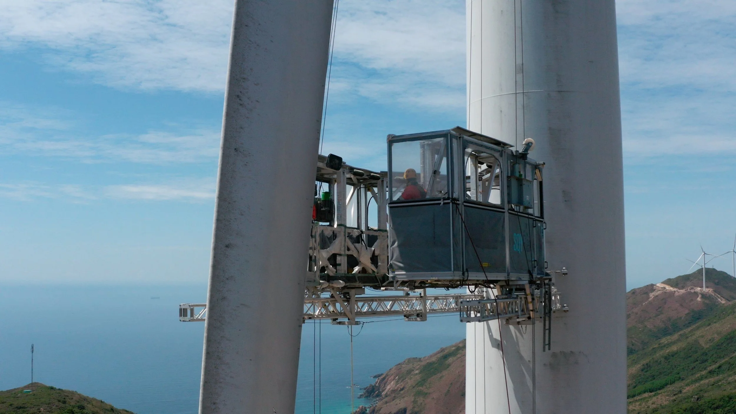 Close-up view of a wind turbine maintenance platform with a worker inside, set against a coastal landscape with wind turbines on distant hills and a blue sky with clouds.