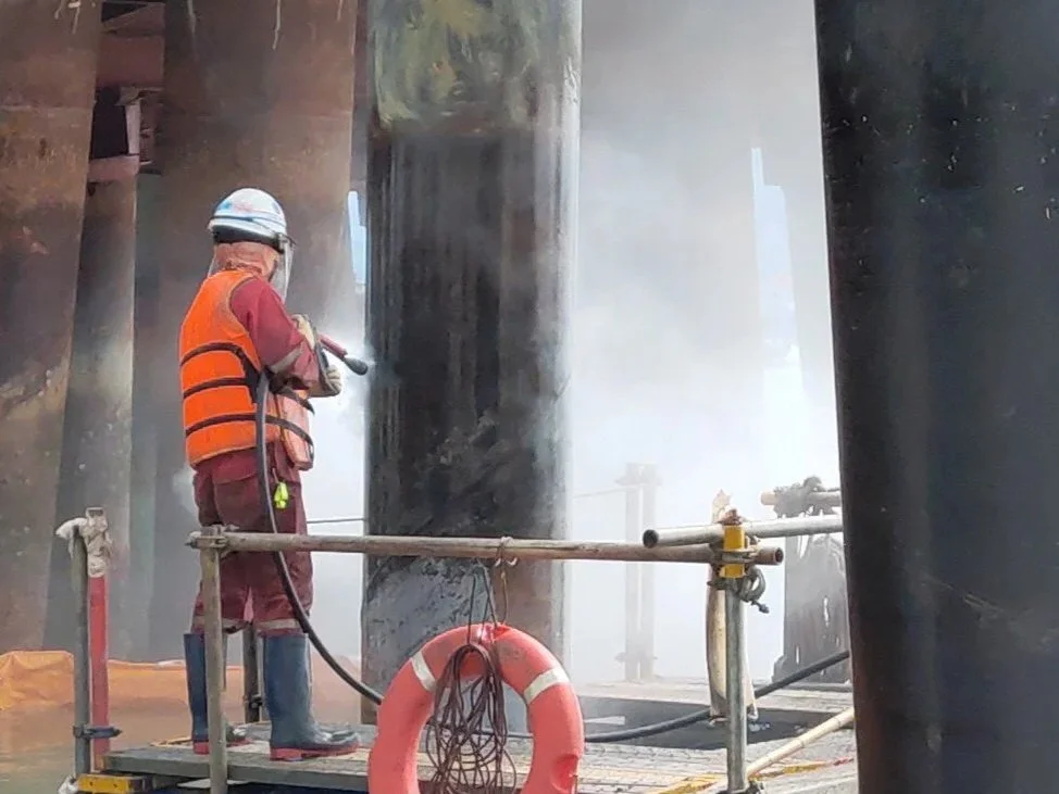 A worker wearing a helmet, orange safety vest, red jacket, and rubber boots is standing on a floating platform under a bridge, using a tool to perform maintenance or inspection on the submerged bridge pilings amidst water spray and mist.