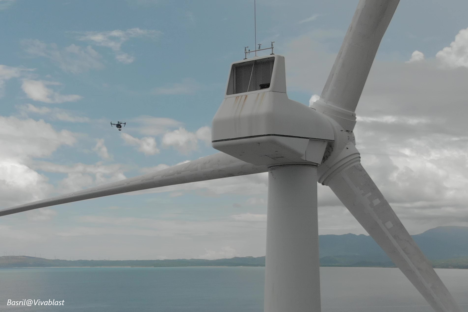Person in high-visibility safety gear operating a drone near a concrete pier with yellow railings, orange traffic cones, and wind turbines in the distance on a clear day.