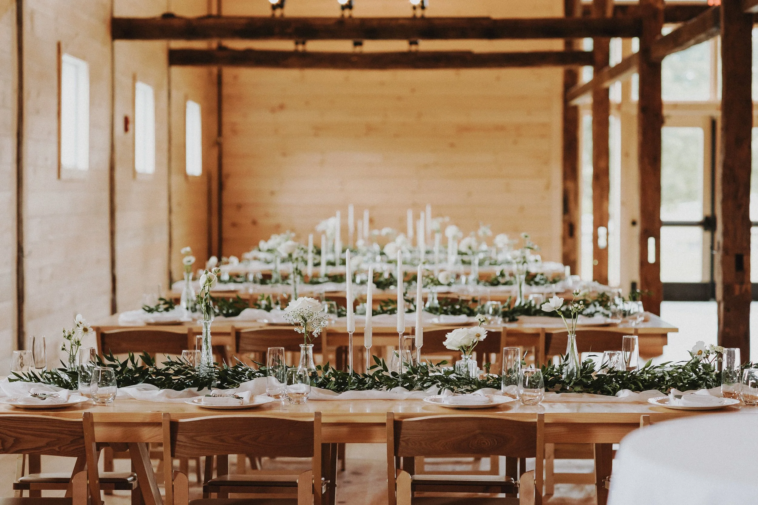 Wedding or event reception setup with long wooden tables, white flowers, candles, and glassware inside a rustic wooden building.