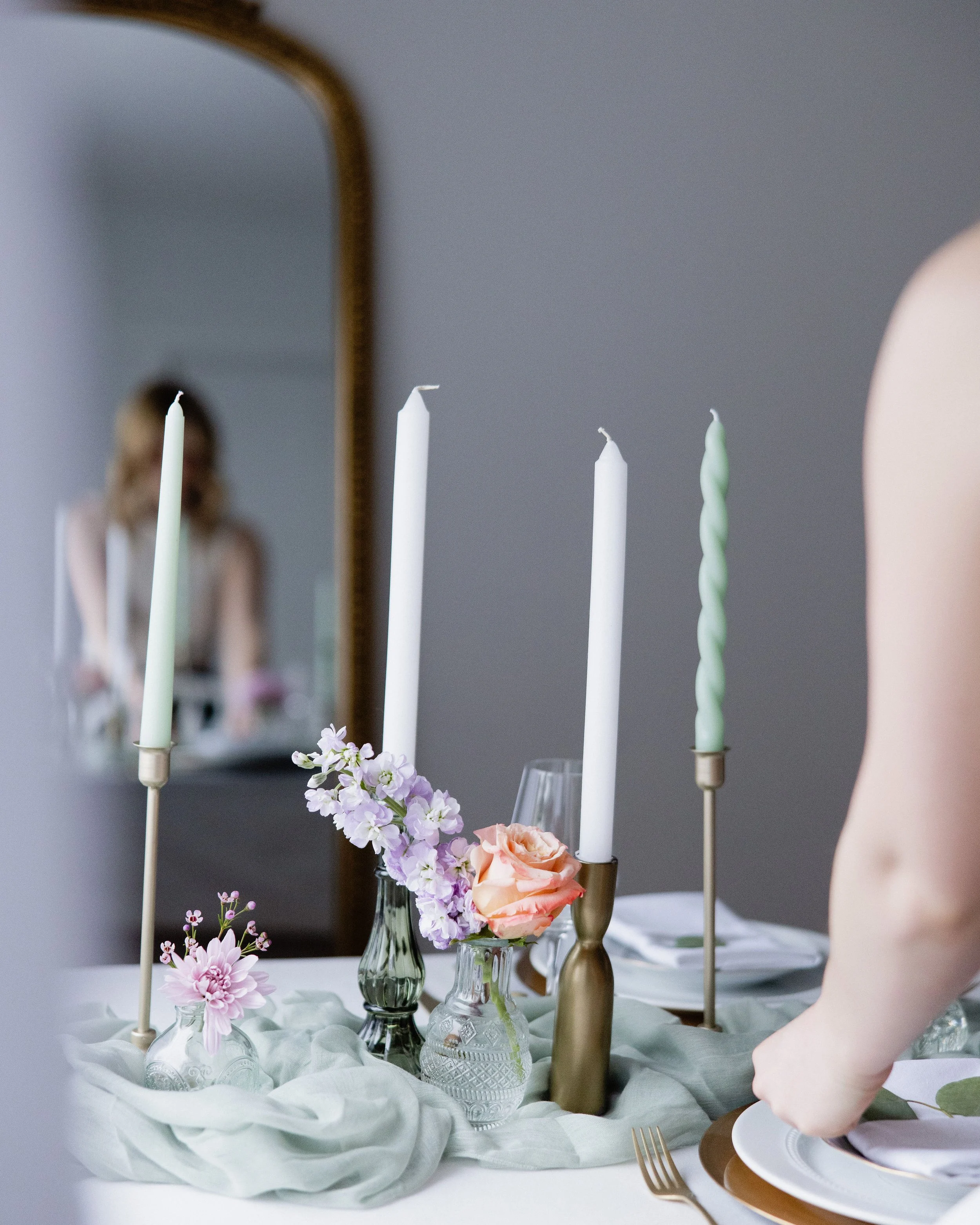 Decorative table setting with pink and purple flowers, four tall candles in gold and glass holders, and a mirror in the background reflecting a woman in a white sash.