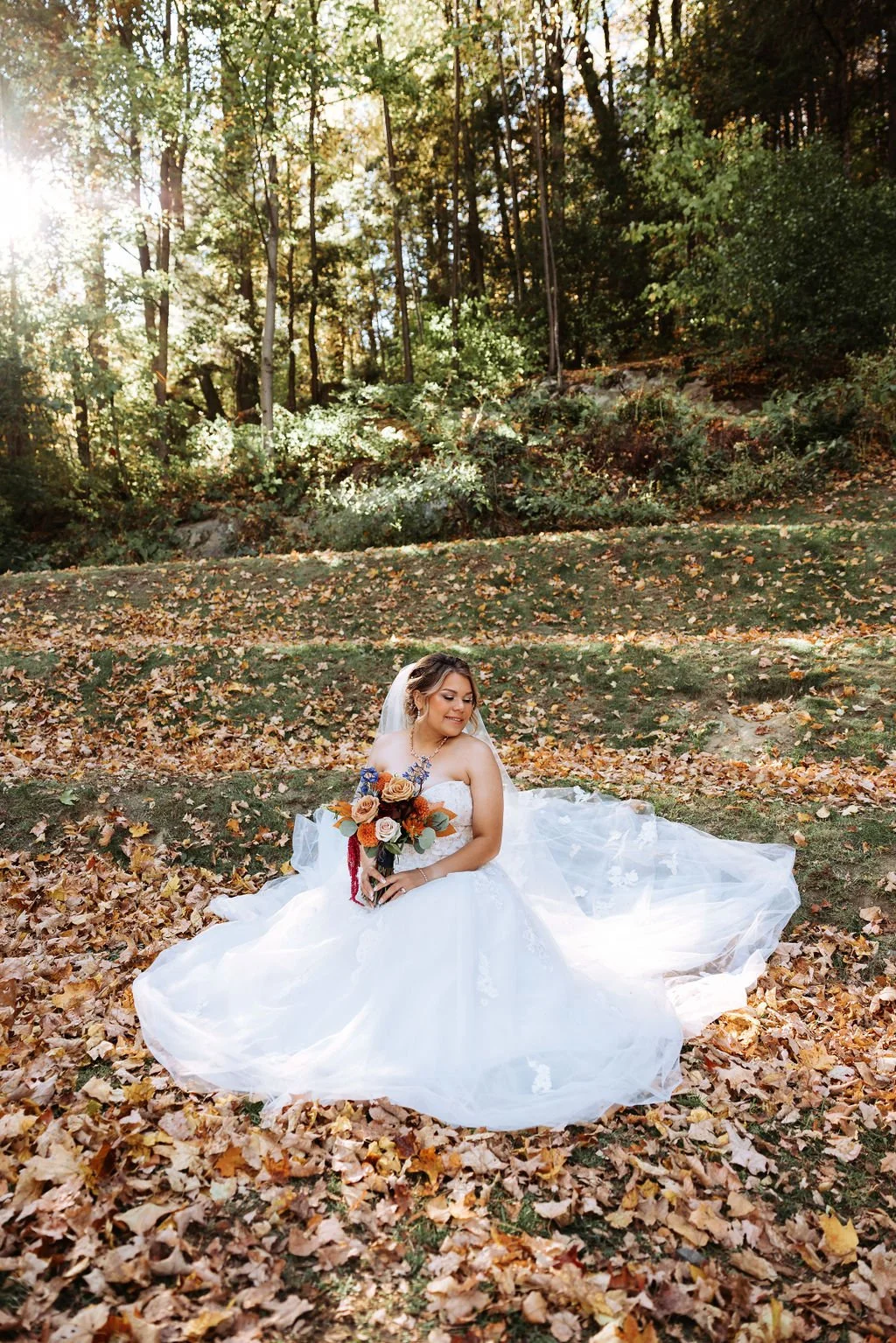 A bride in a wedding dress sitting on fallen leaves in a wooded outdoor setting, holding a bouquet of orange, pink, and cream roses, with sunlight filtering through trees in the background.