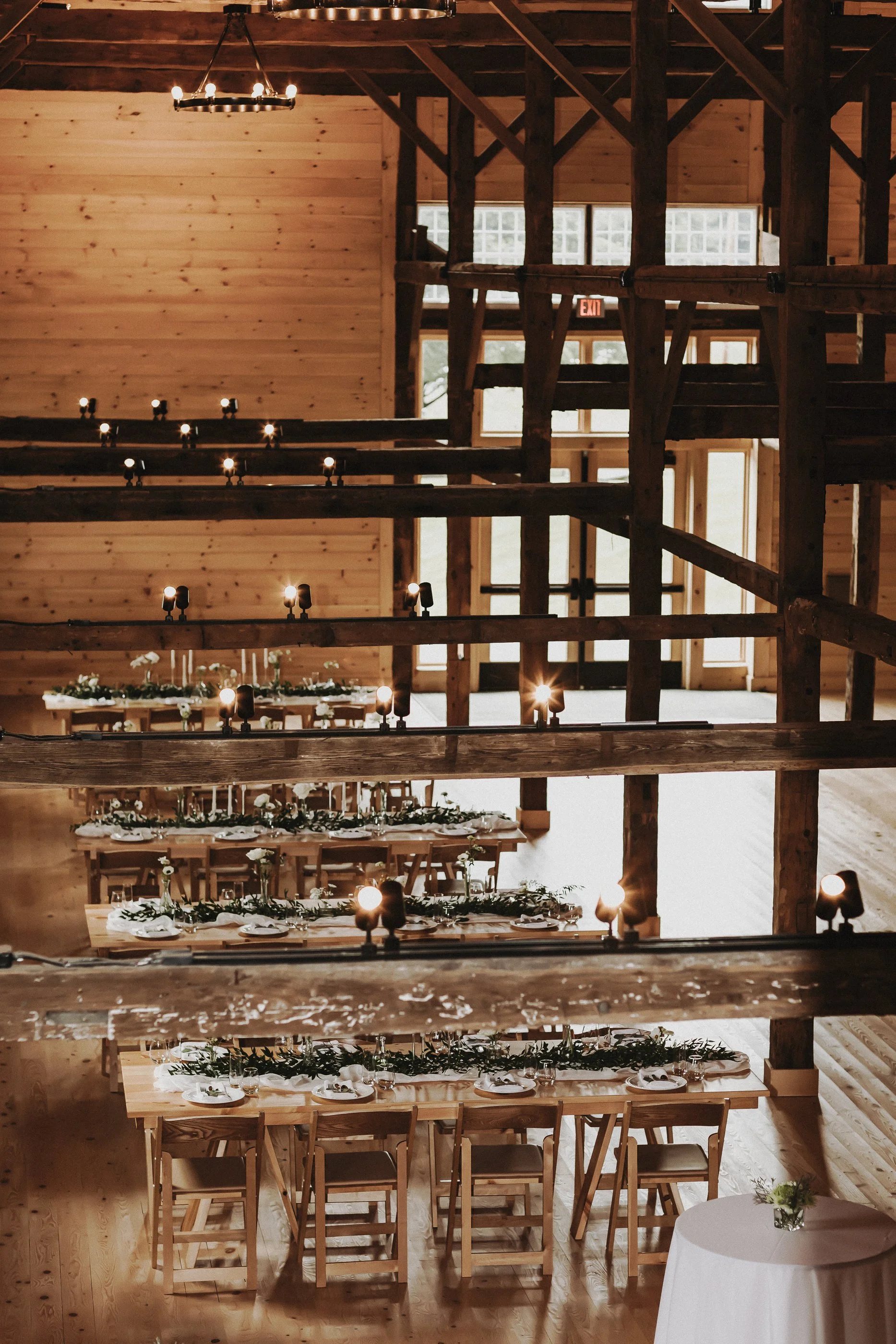 A rustic wooden event space with long tables decorated with greenery and white flowers, set for a meal, illuminated by hanging lights. The space features a high ceiling with exposed wooden beams and natural light coming through windows and a glass door.