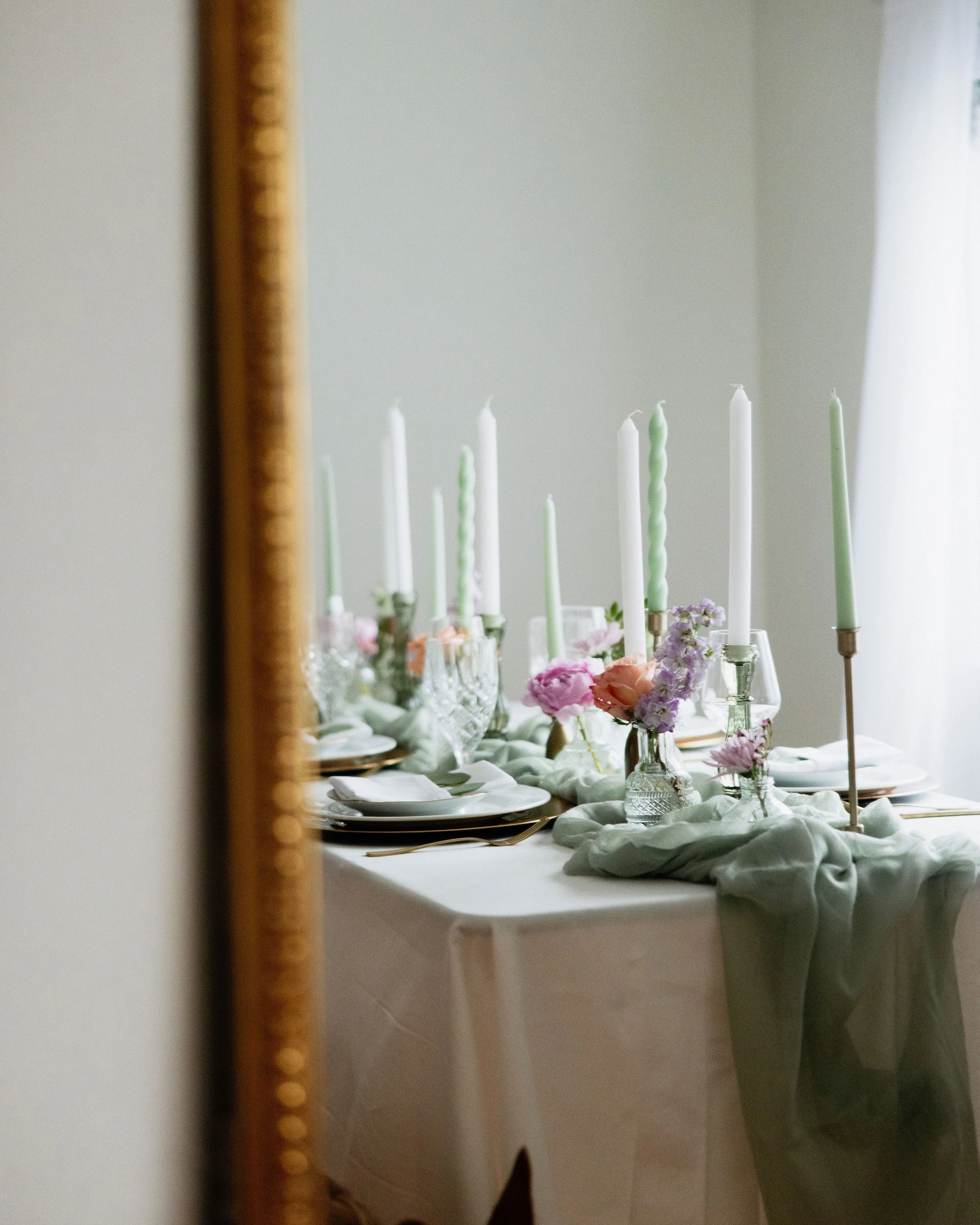 A decorated dining table with floral centerpieces, pastel-colored candles, and neatly arranged tableware seen through a mirror with a gold frame.