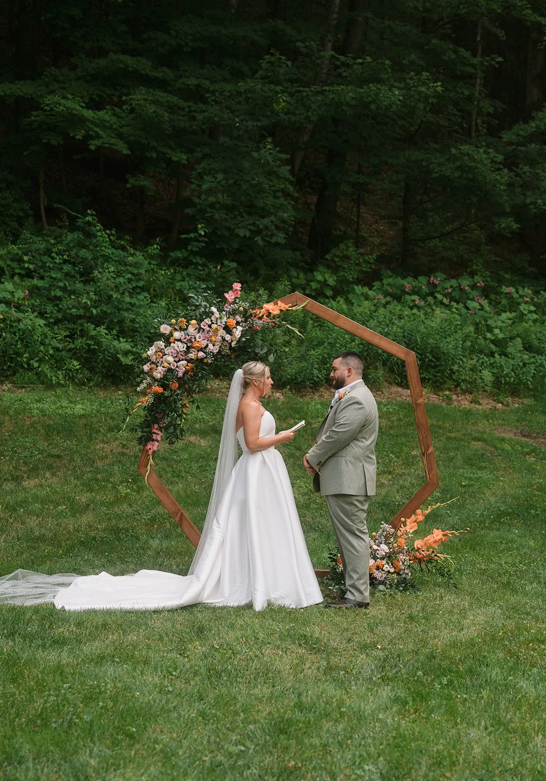 A bride and groom standing in front of a floral arch during their outdoor wedding ceremony on a grassy lawn with a wooded background.