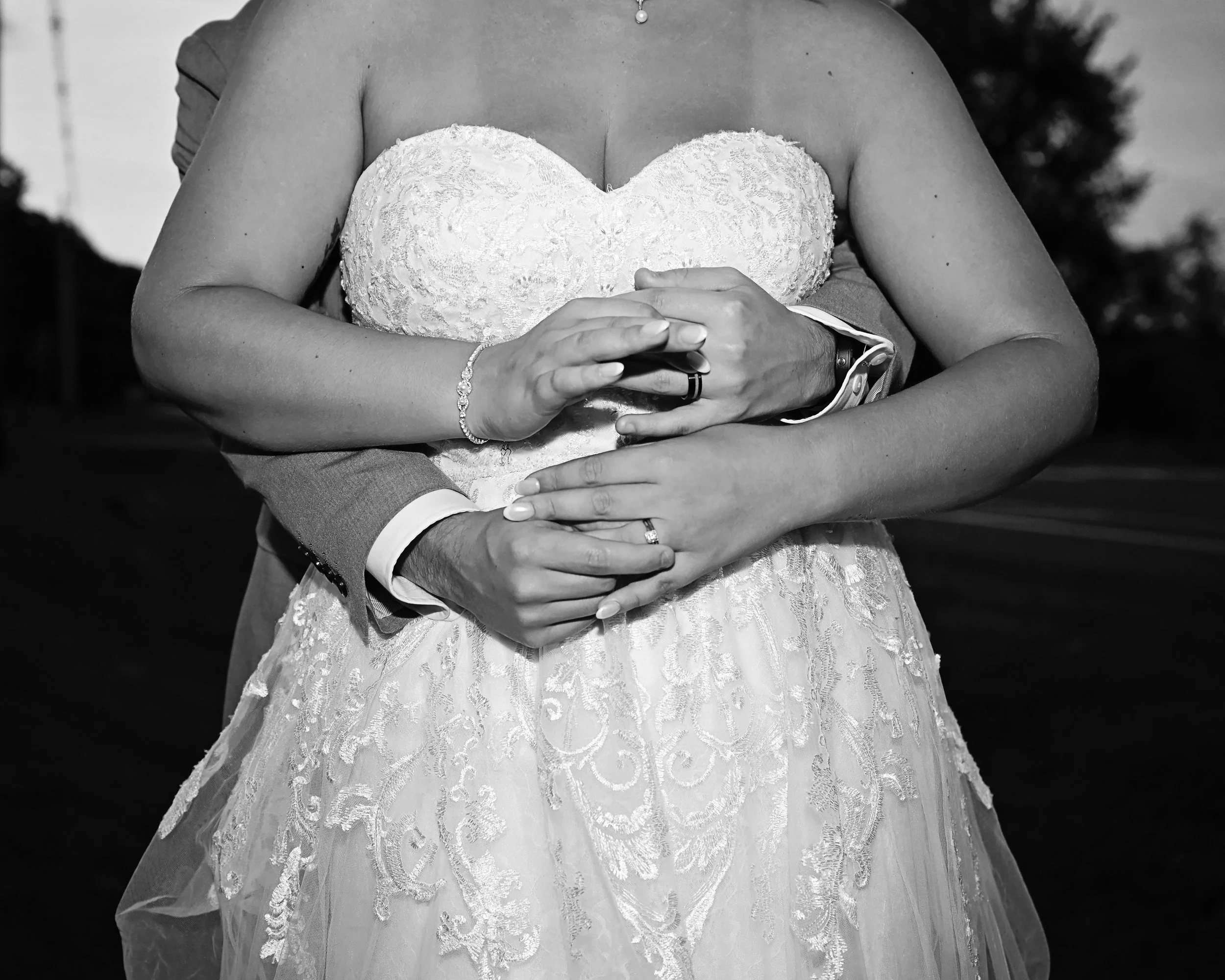 Black and white photo of a bride and groom holding hands, with the groom's arms around the bride, showing rings and wedding dress.