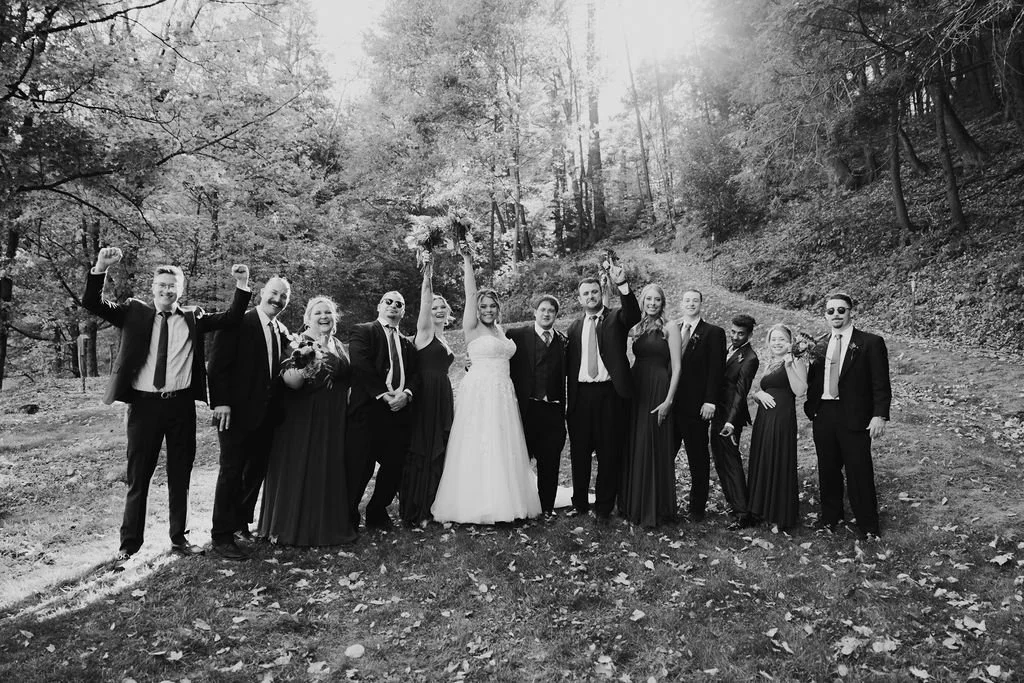 Black and white photo of a wedding party outdoors in a wooded area, with the bride and groom in the center and friends and family celebrating around them.