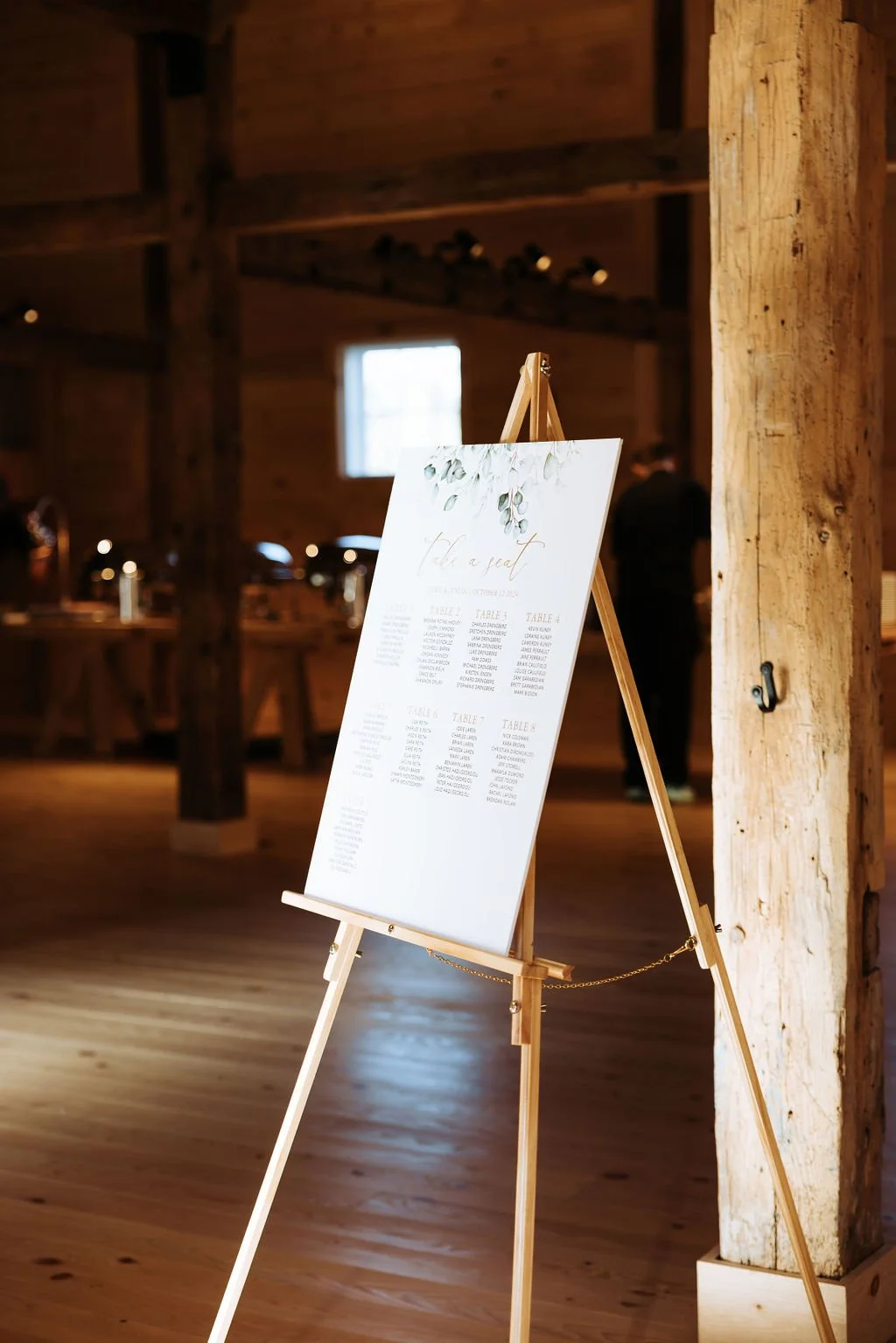 Wedding seating chart on a white board with floral decoration, displayed on a wooden easel in a rustic venue with wood beams and wooden walls.