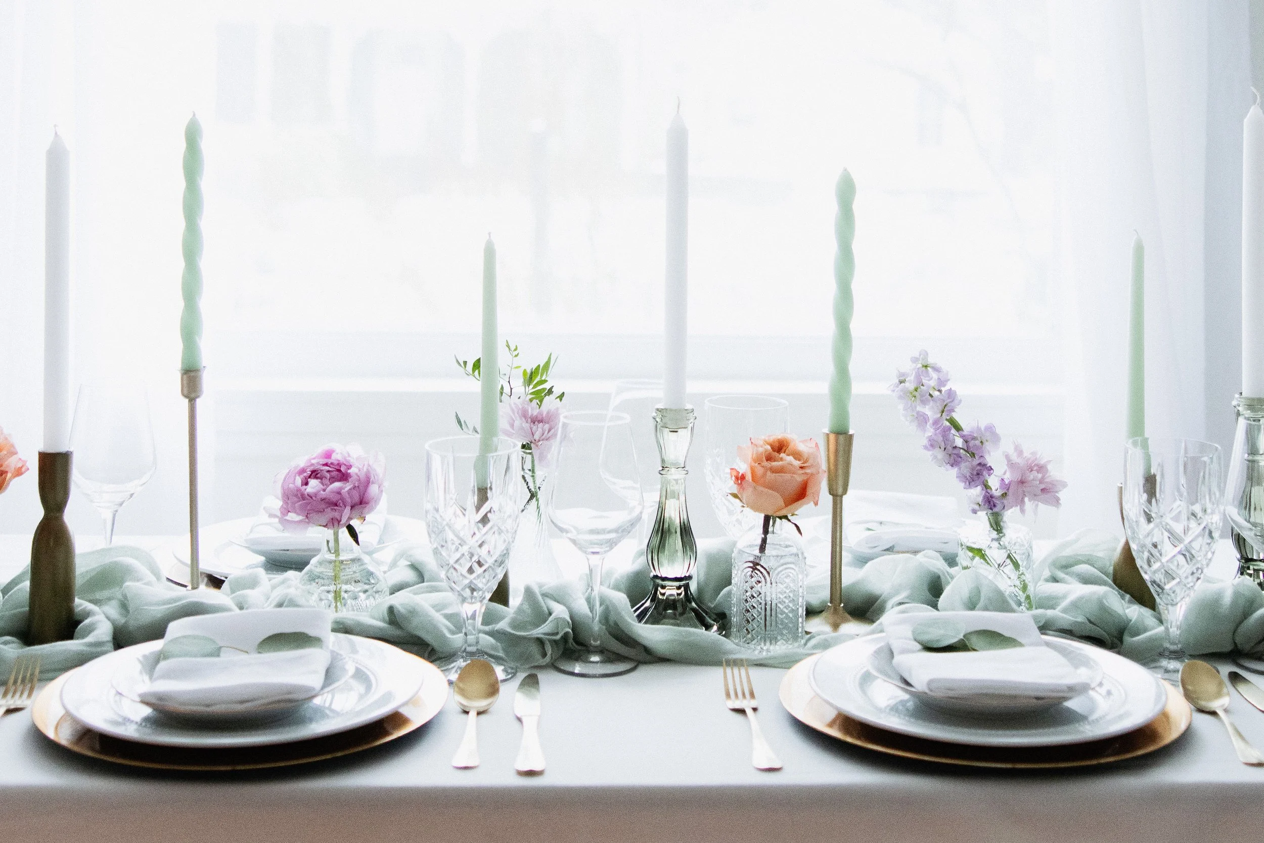 Elegant table setting with white plates, gold-rimmed chargers, cutlery, crystal glasses, and floral centerpieces with pink, purple, and peach flowers, set beneath pastel-colored candles in a bright room.
