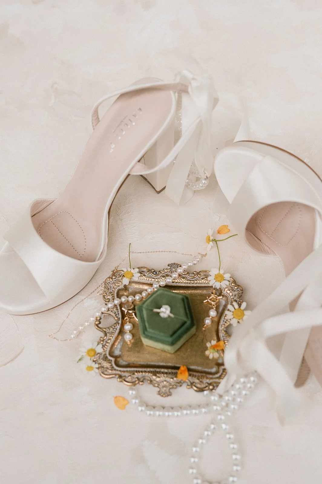 A pair of elegant white high heel shoes with ankle straps, a vintage jewelry tray with pearl necklaces and earrings, a small green ring box, and scattered white and yellow flower petals on a light-colored surface.