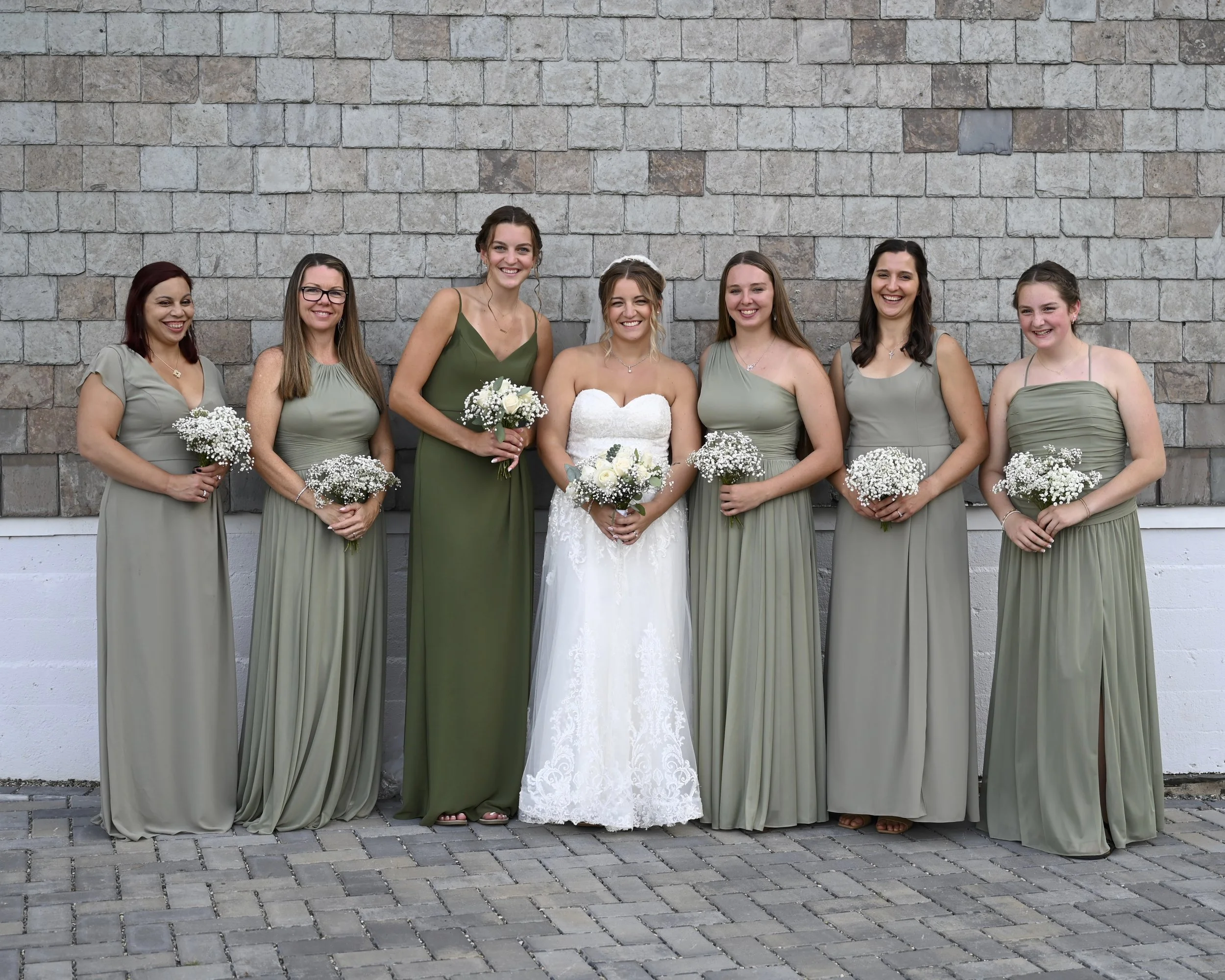 Group of seven women at a wedding, with the bride in a white gown and six bridesmaids in green dresses, all holding small bouquets of white flowers, standing against a brick wall background.