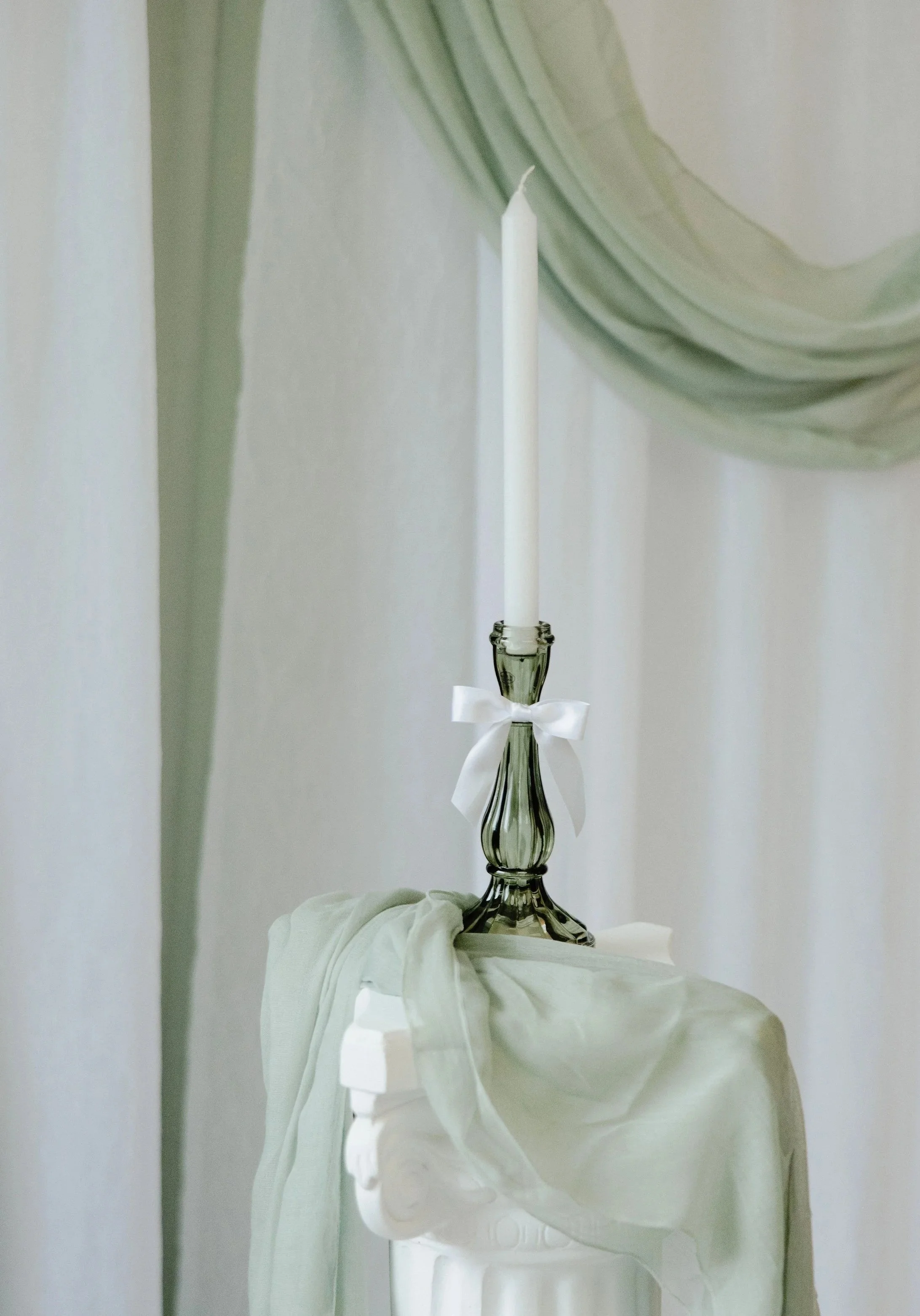 A white candle in a glass candle holder with a white ribbon, placed on a decorative white pedestal draped with a light fabric.
