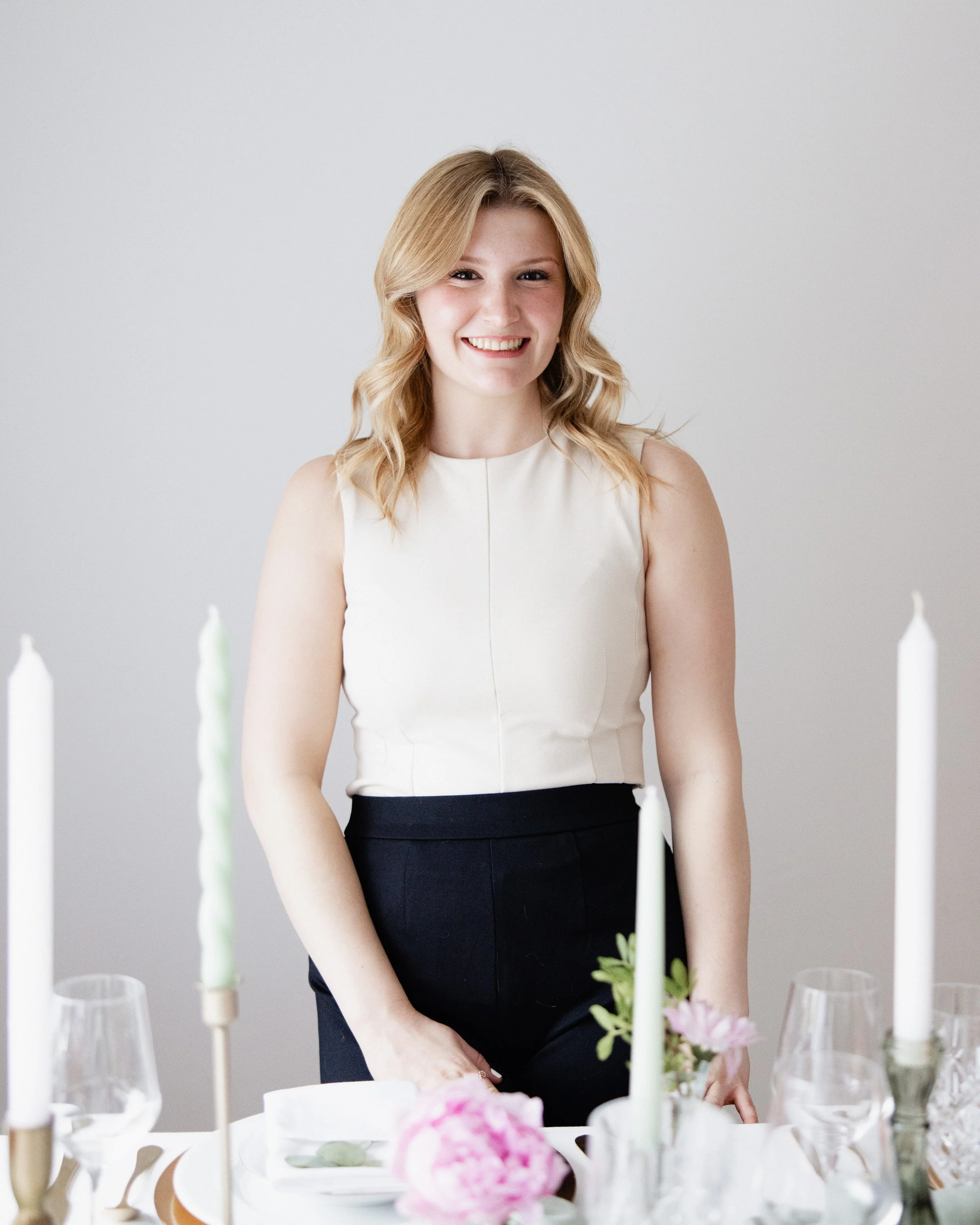 A woman with blonde, wavy hair smiling at a dinner table with candles and pink flowers.