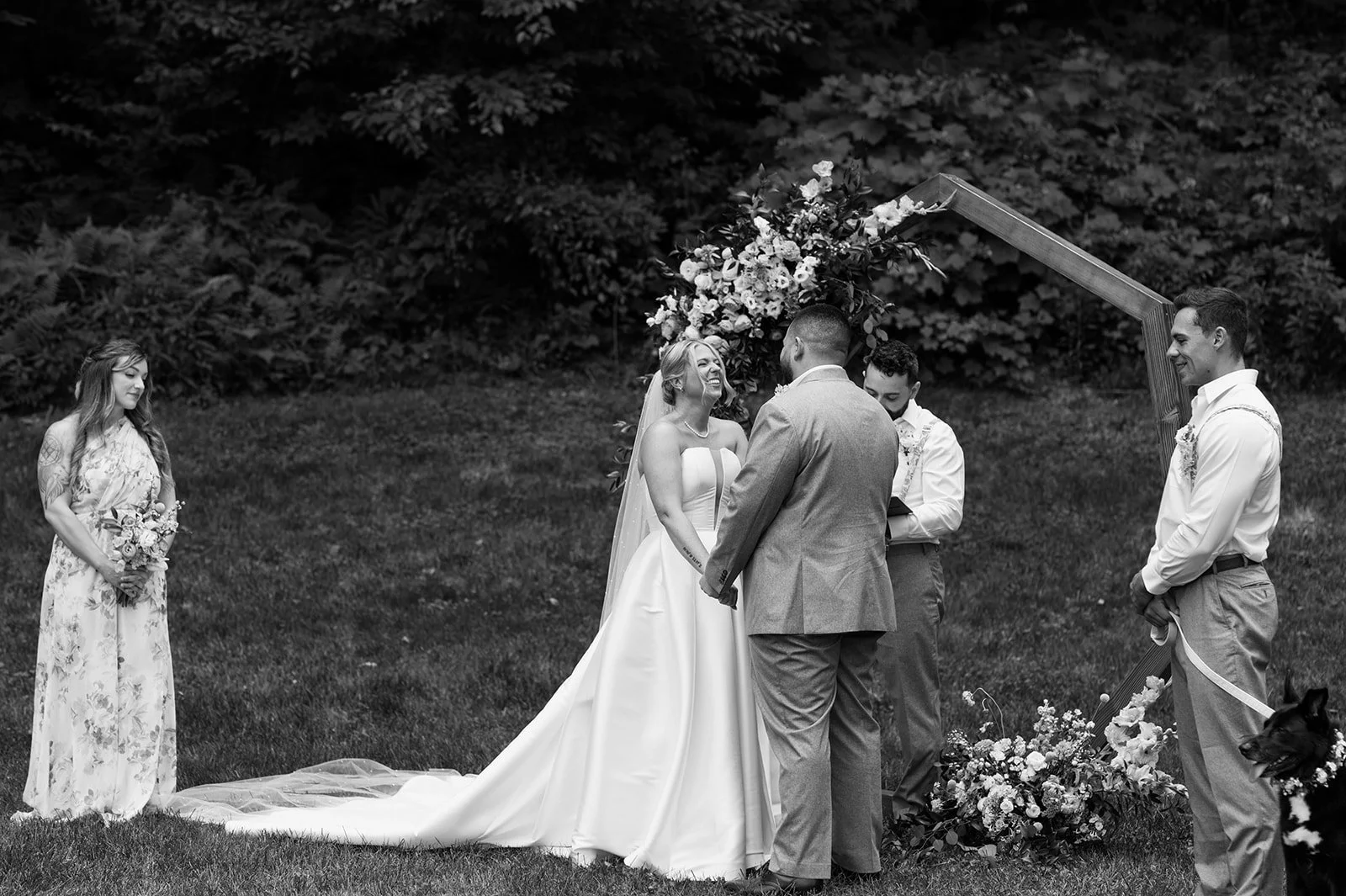 Black and white photo of a wedding ceremony outdoors with a bride, groom, officiant, bridesmaid, and groomsman under a wooden arch decorated with flowers, on grass with trees in the background.