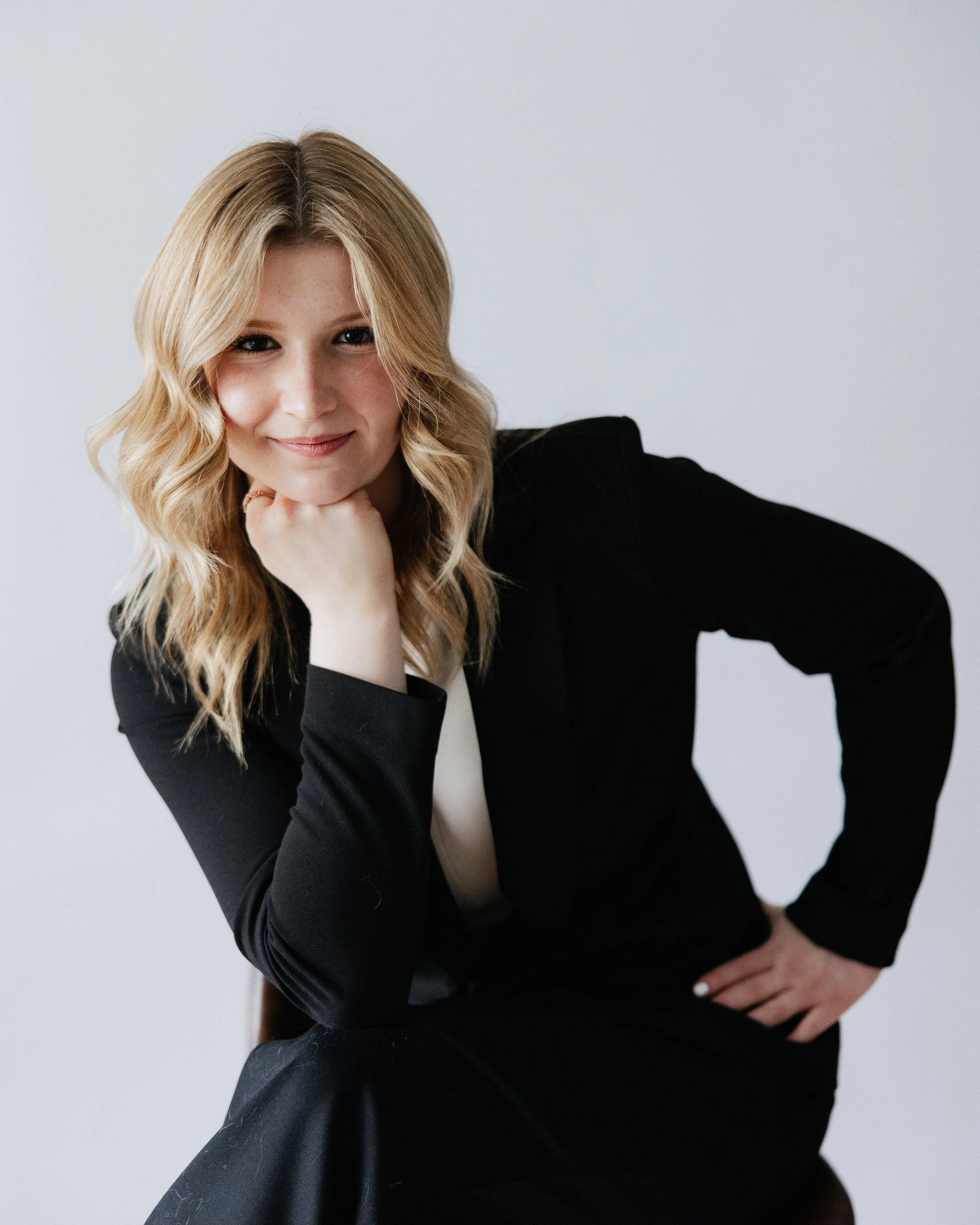 A woman with blonde wavy hair, wearing a black blazer, sitting with her chin resting on her hand, smiling at the camera against a light gray background.