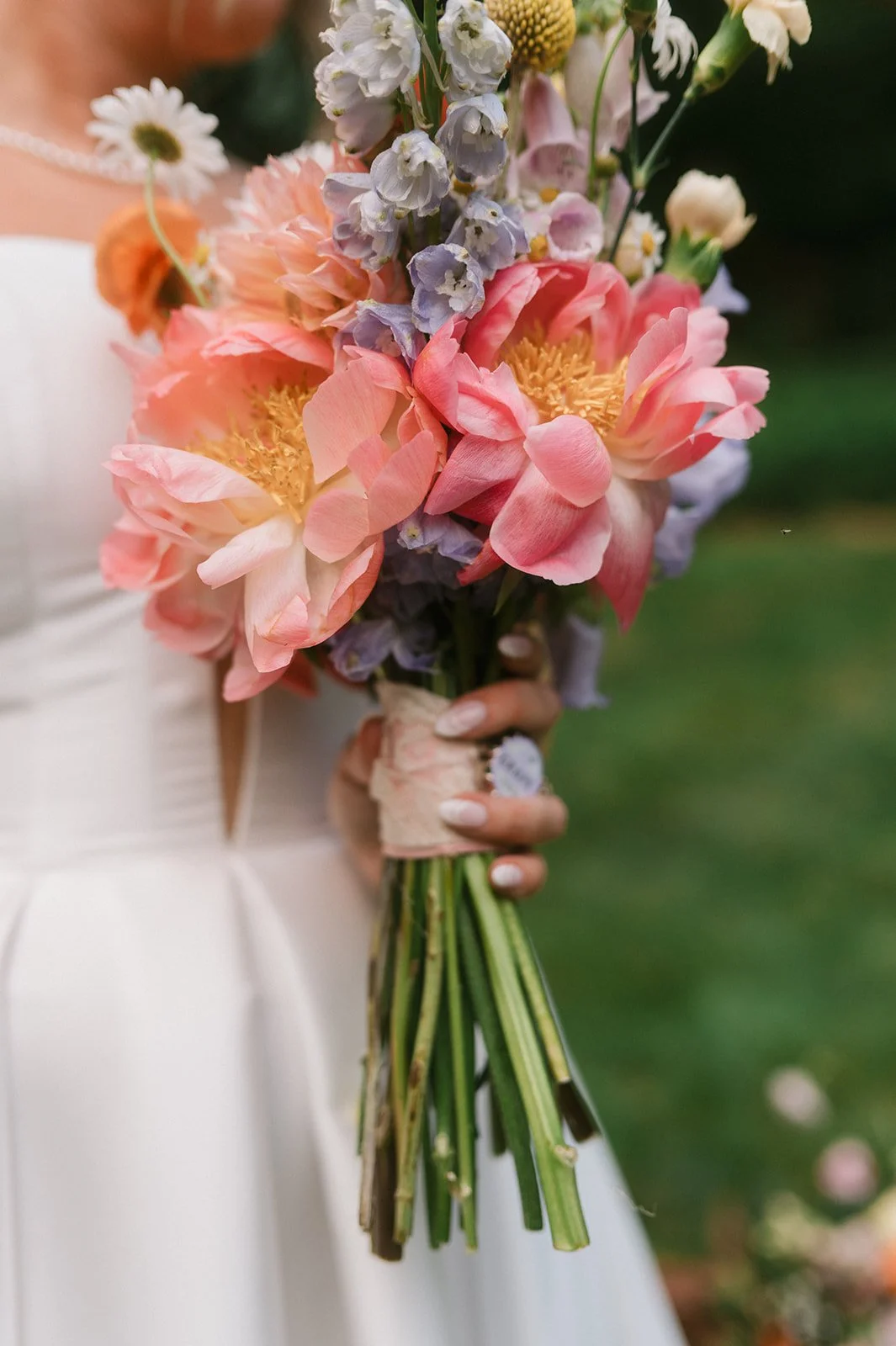 A bride holding a bouquet of pink, purple, and white flowers, with the focus on the flowers and her white dress visible in the background.
