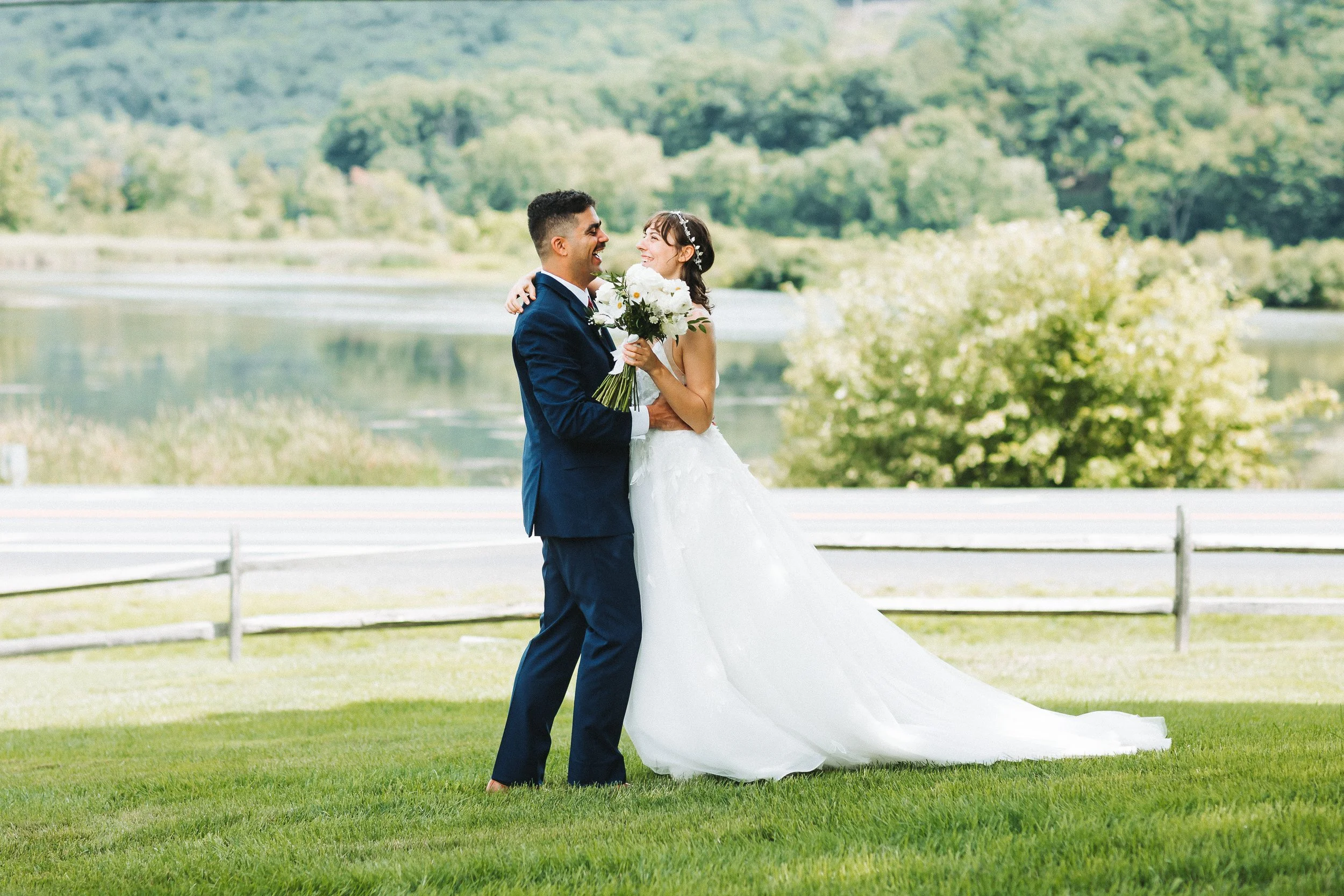 A newlywed couple in wedding attire standing on grass near a road, with a scenic lake and trees in the background. The bride is holding a bouquet and smiling at the groom, who is dressed in a navy suit.