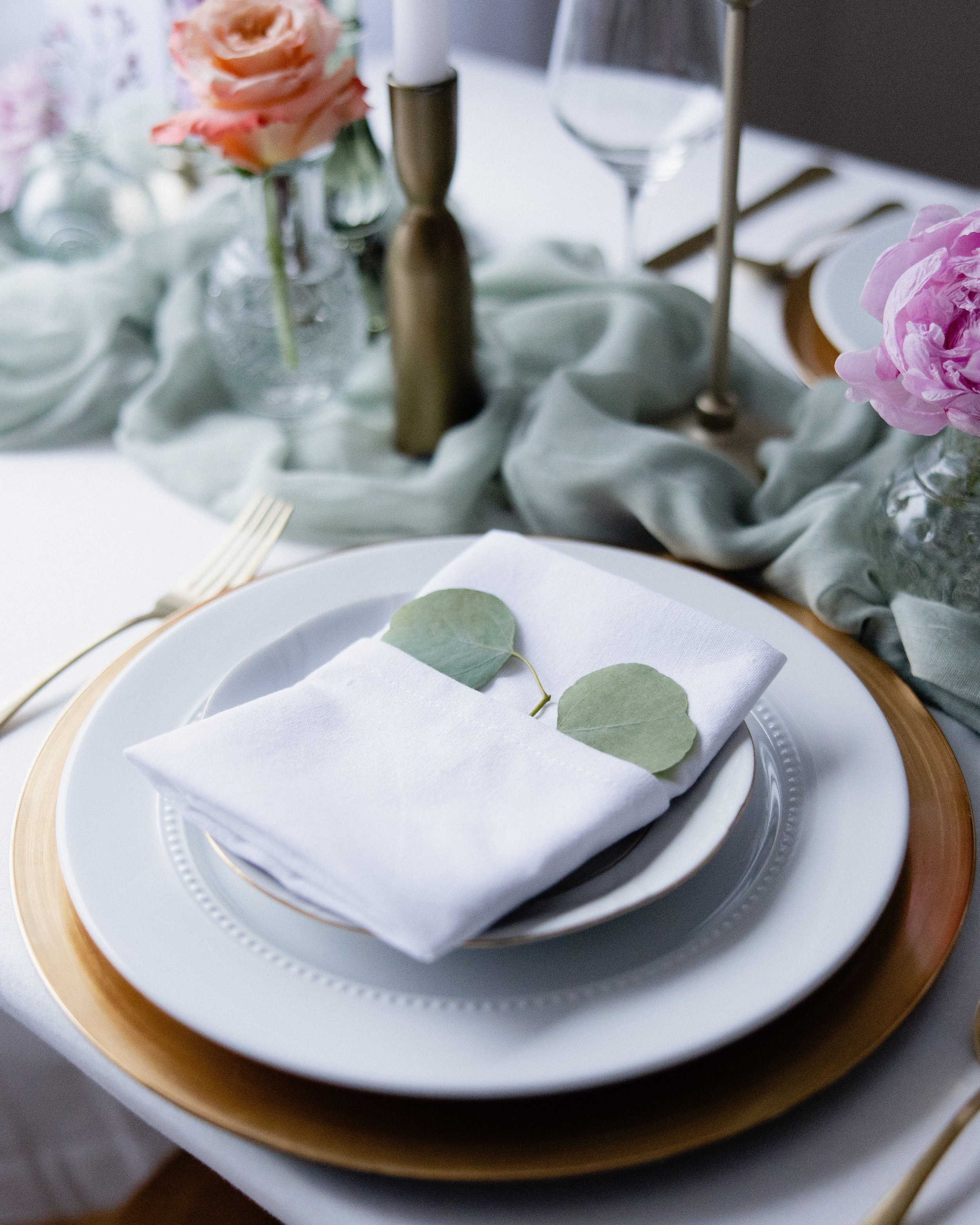 A formal place setting with a white napkin and green leaves on top of a white plate, with other plates, gold flatware, and a decorated table with flowers and candles in the background.