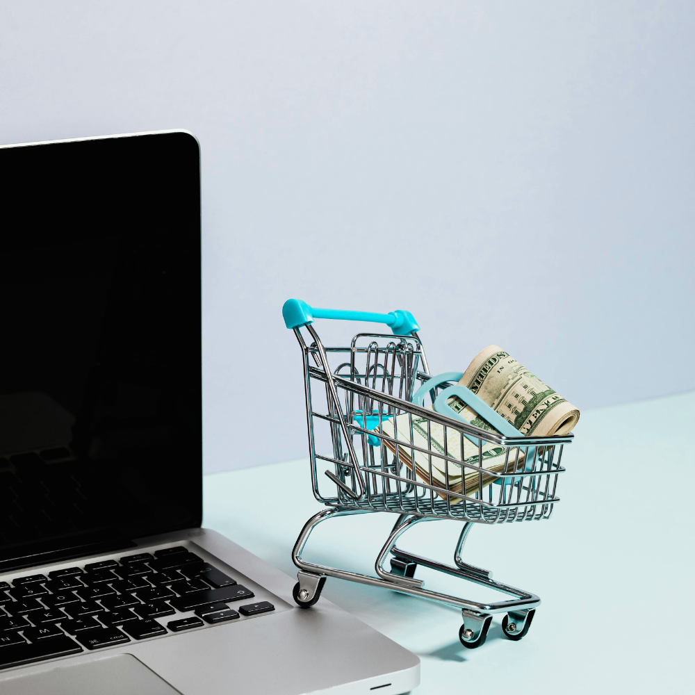 Miniature shopping cart with rolled US dollar bills next to a laptop on a white surface.