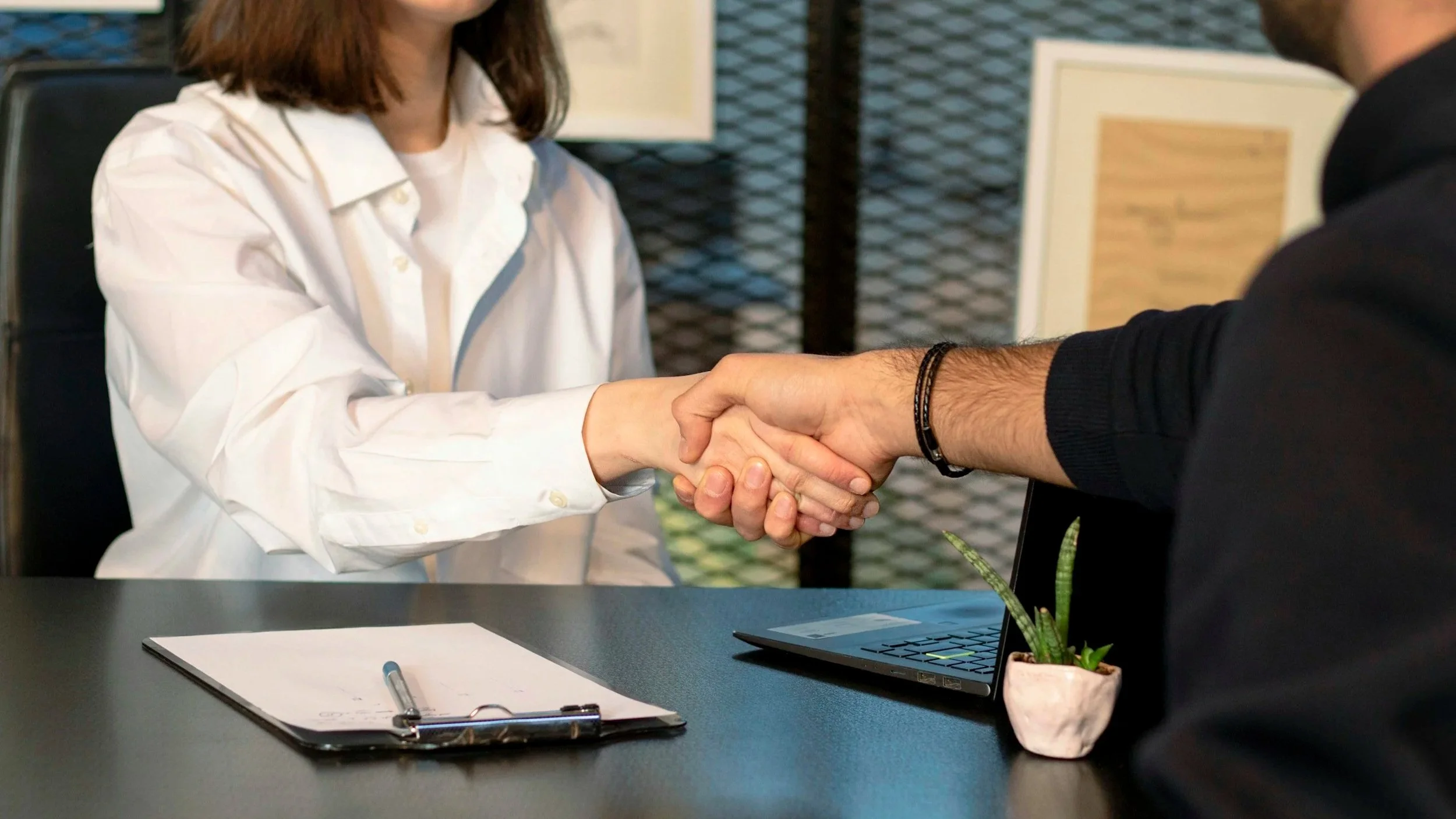 A woman in a white shirt shaking hands with a man in a black shirt at a table in an office setting, with a clipboard, pen, and laptop on the table, and framed pictures on the wall in the background.