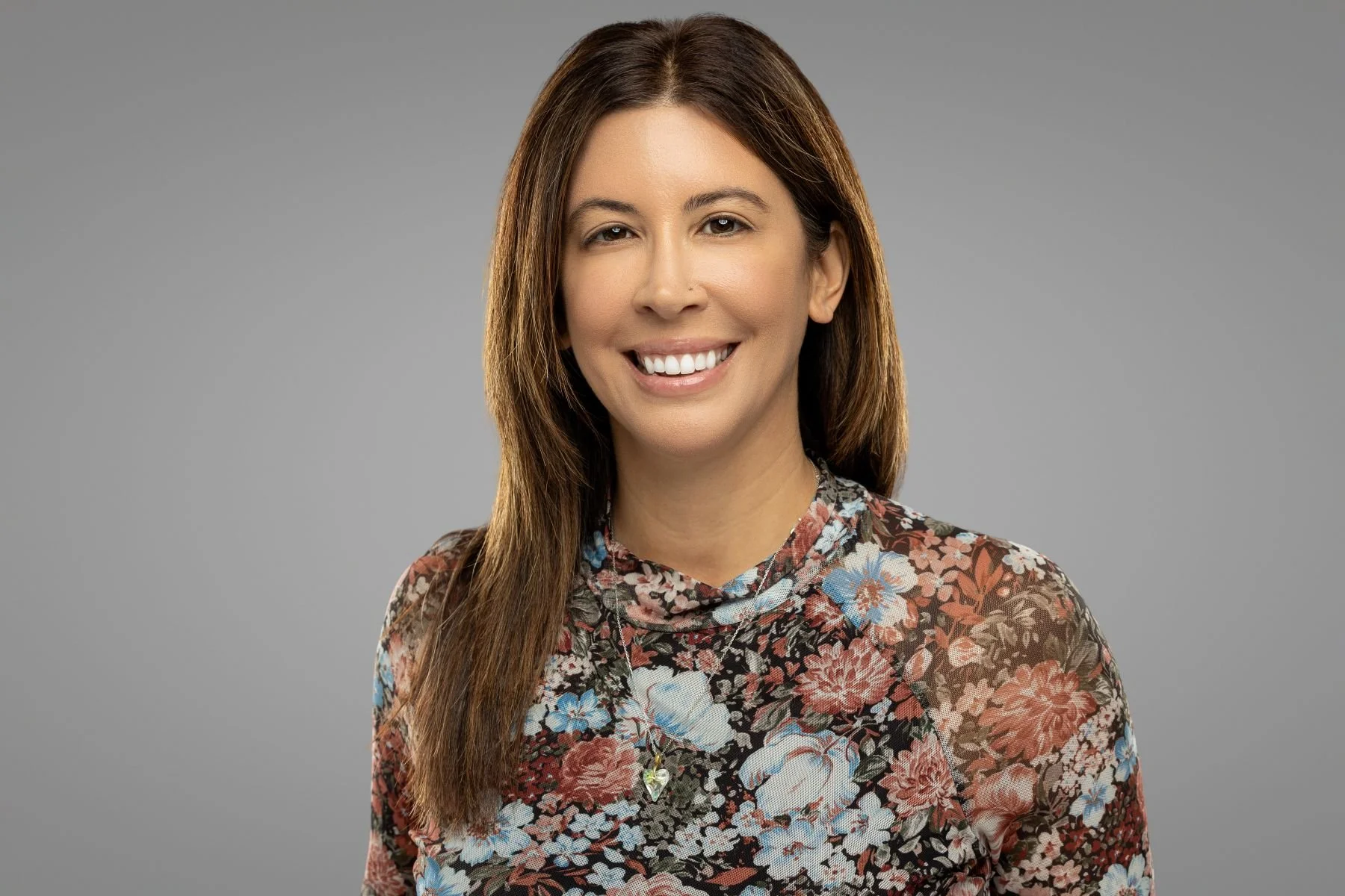 A smiling woman with long brown hair wearing a floral patterned top against a gray background.