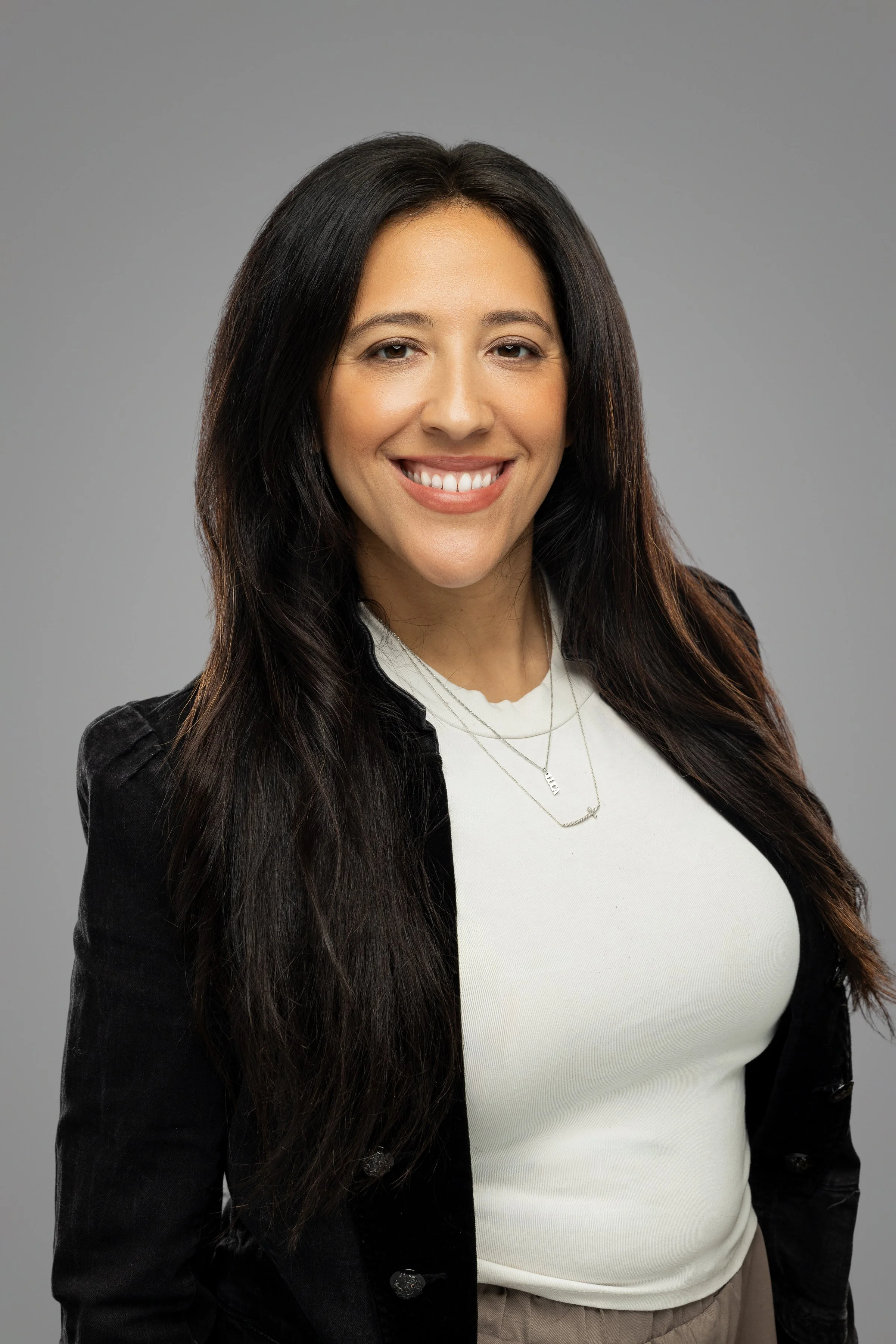 A woman with long, dark hair smiling, wearing a white top, black jacket, and layered necklaces, against a grey background.
