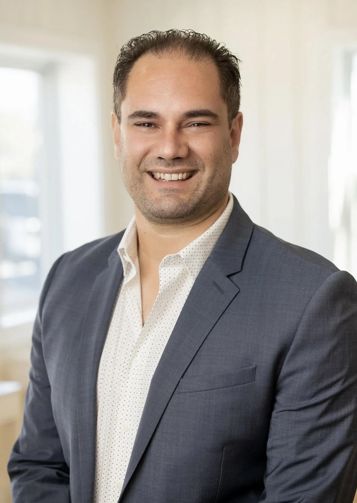 A man with dark hair in a business suit smiling in an indoor setting.