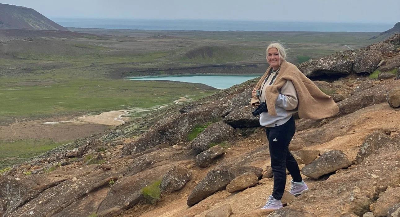 A woman standing with a geothermal hot spring visible in the distance at Krýsuvík Geothermal Area.