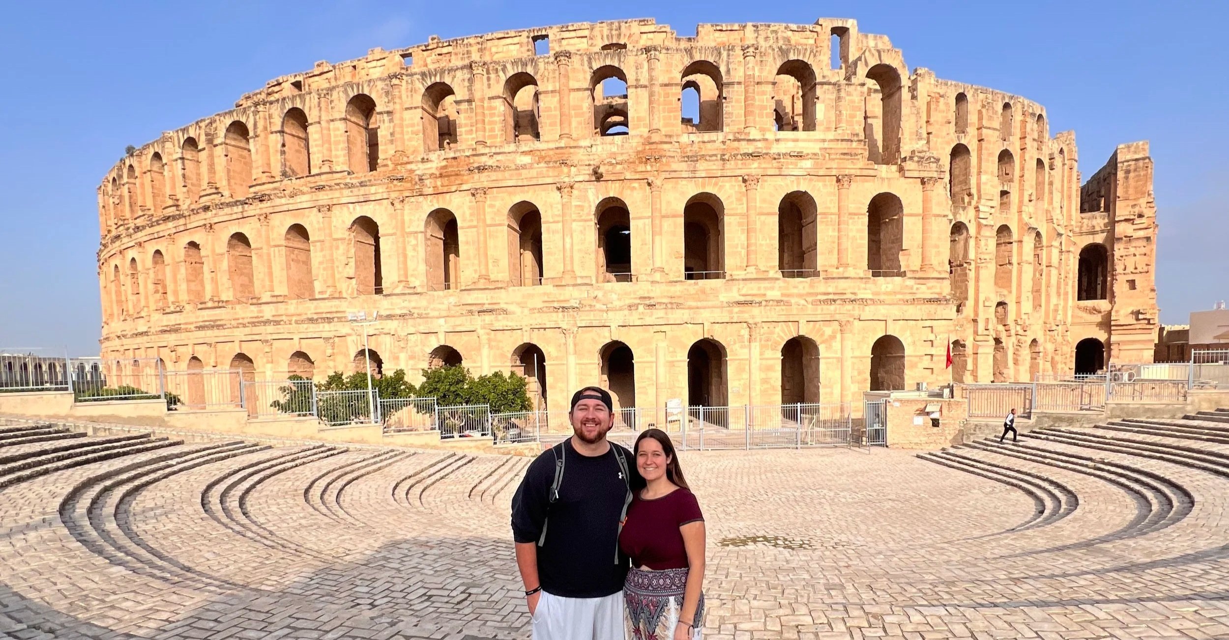 A couple standing in front of the El Jem Amphitheater in Tunisia in the morning, with the historic Roman ruins illuminated by sunlight.