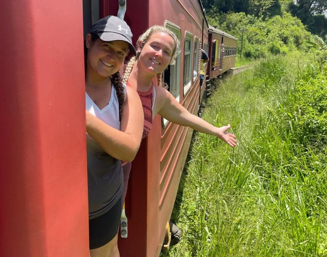 Two women smiling and leaning out of a moving train in Sri Lanka, enjoying the ride.