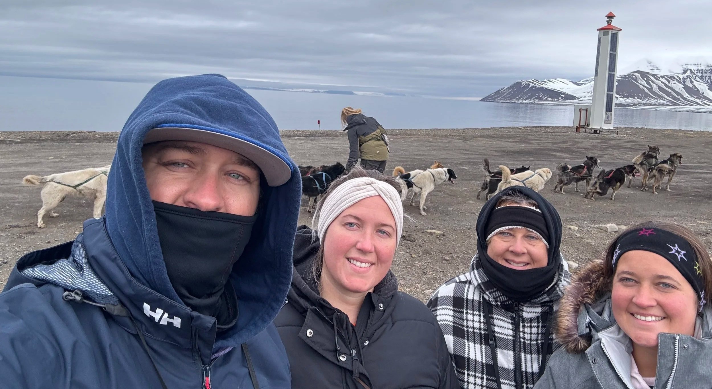 Three women and one man smiling in front of a dog sled team pulling a wagon during summer, Arctic tundra landscape in the background