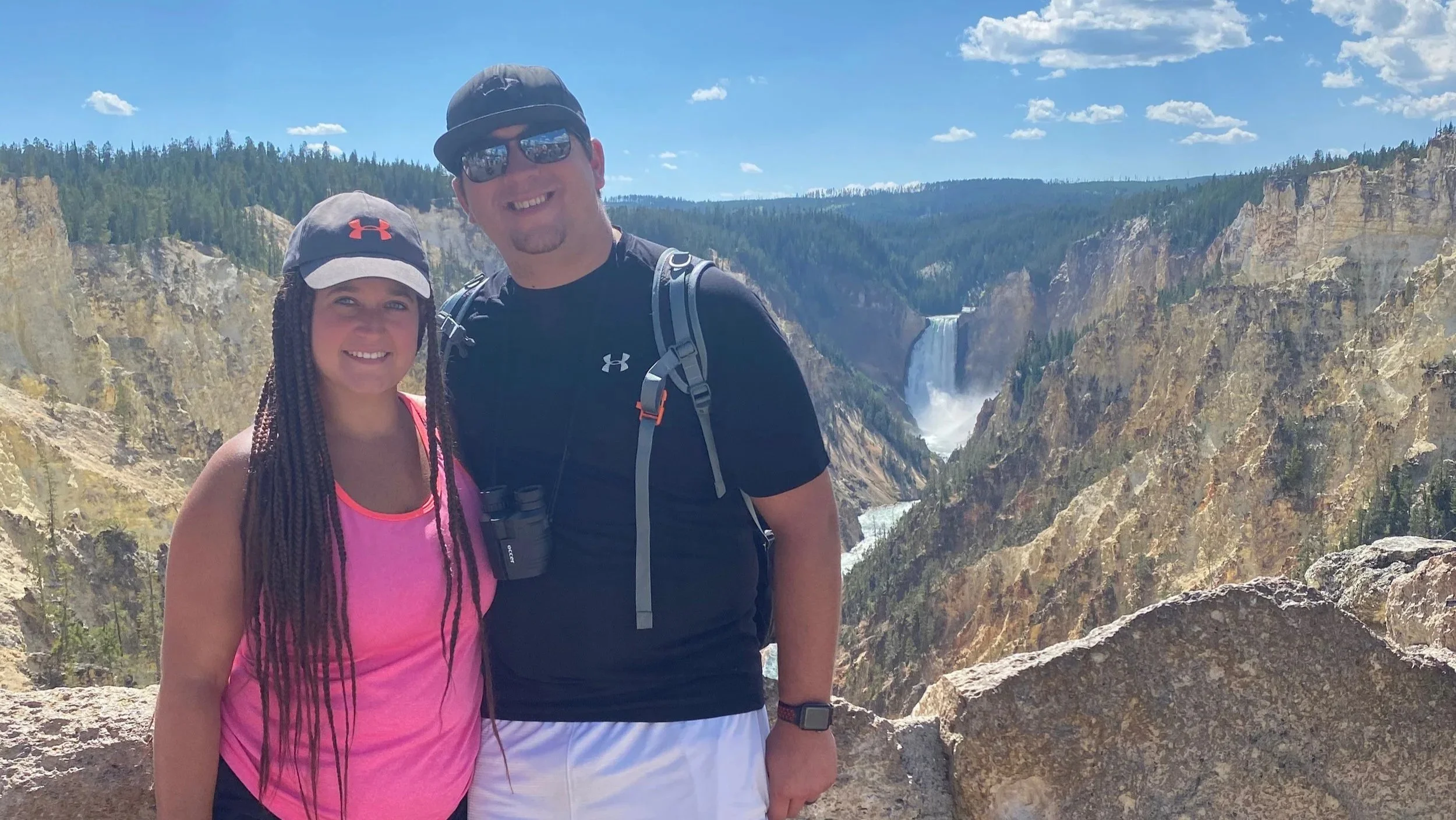 A couple smiling in front of the cascading waterfalls at Inspiration Point, surrounded by lush greenery and rocky cliffs.