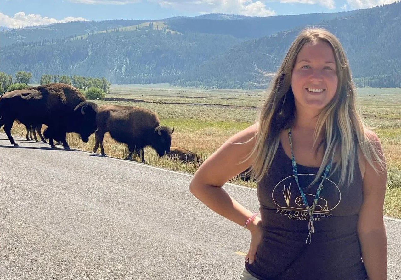 A woman smiling with a bison standing nearby in a grassy, natural landscape in Hayden Valley.