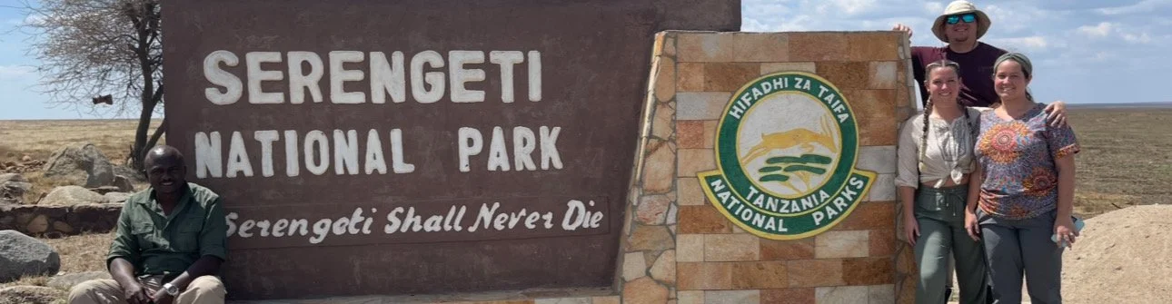 Three people with their safari guide posing in front of the Serengeti National Park sign in Tanzania.