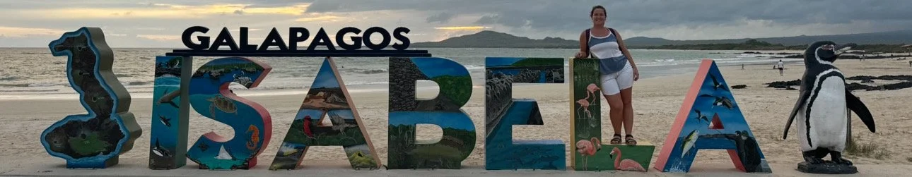 A young woman standing in front of the Isabela Island sign with the beach behind in the Galápagos Islands.