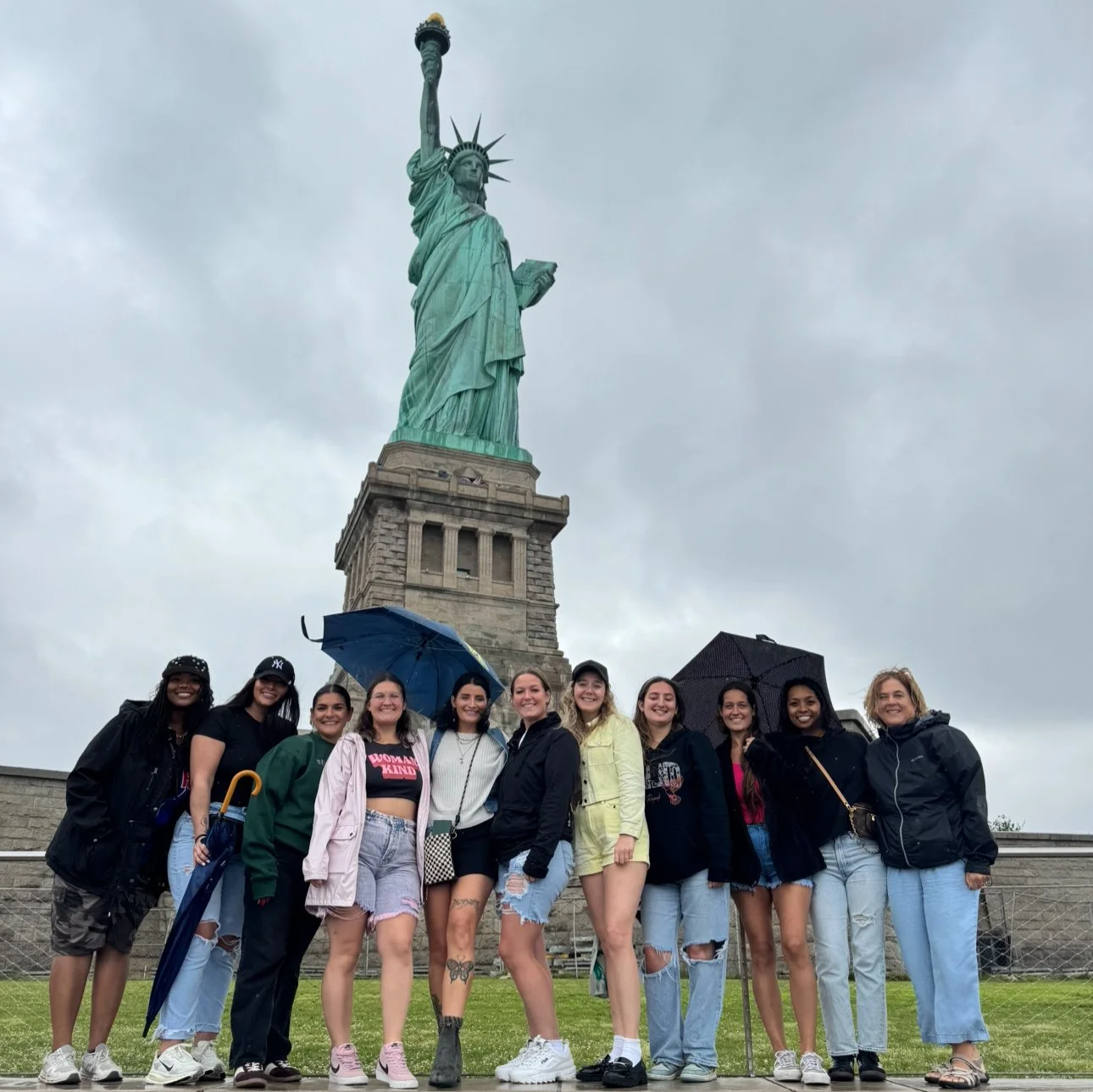 Eleven girls smiling together in front of the Statue of Liberty in New York City.