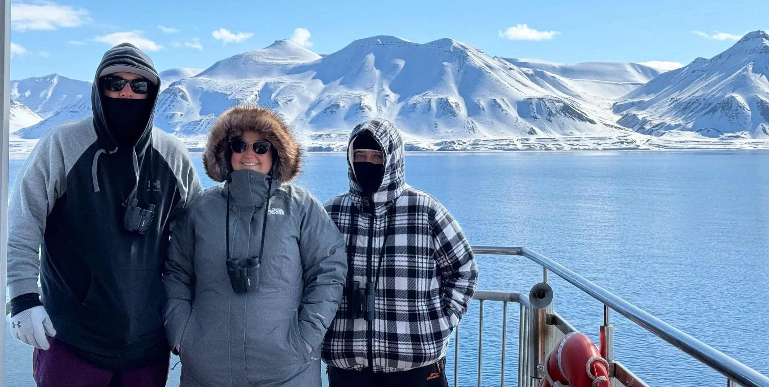 Two women and a man bundled up standing on a boat in the Arctic, snowy mountains rising in the background over icy waters
