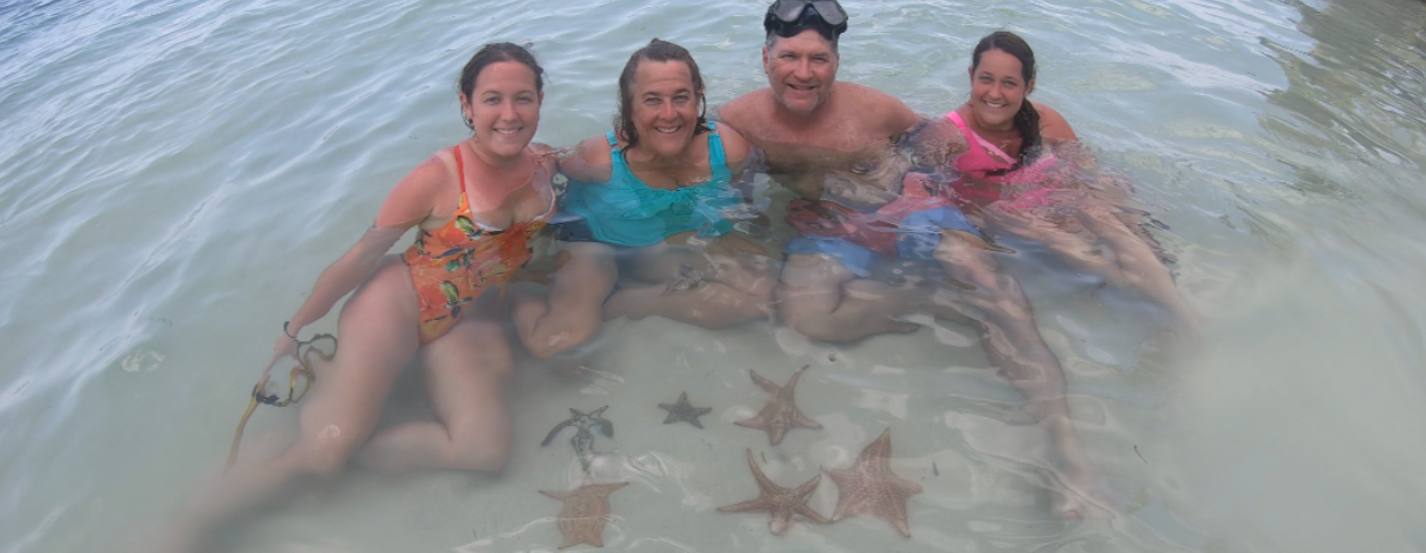 A family sitting in shallow, clear water with several starfish visible in front of them.