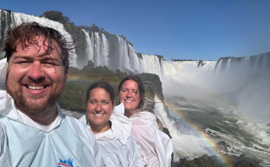Three people walking on the boardwalks at Iguazu Falls, Brazil, with waterfalls visible in the background.